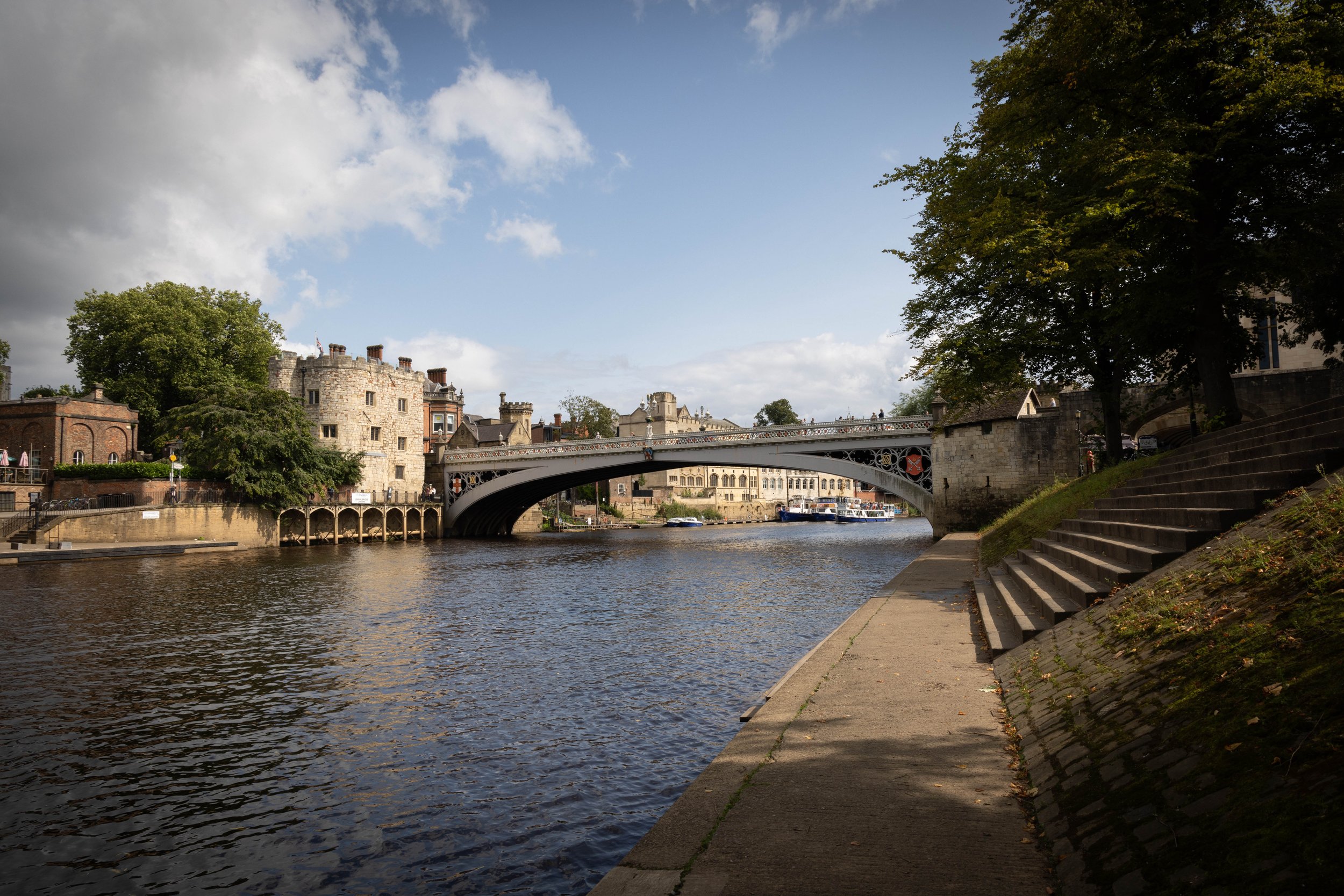 View of a historic stone castle-like building with multiple chimneys, a bridge crossing the river, boats on the water, and trees along the riverbank on a partly cloudy day.