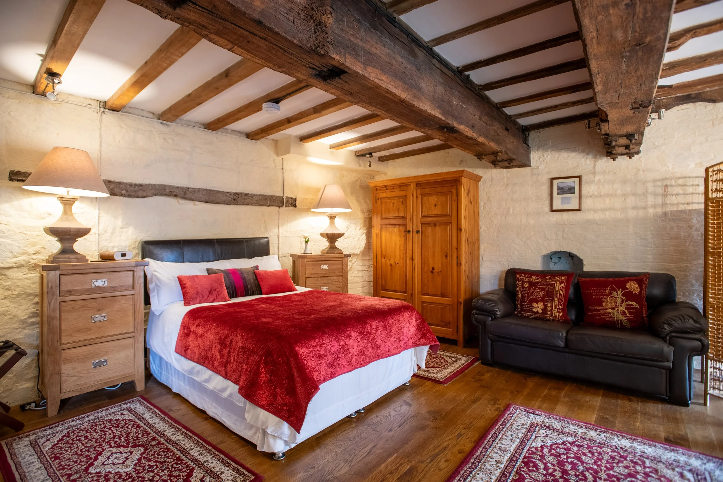 A cozy bedroom with exposed wooden ceiling beams and a brick and plaster wall. It features a bed with red bedding and multiple pillows, two matching wooden nightstands with lamps, a black leather sofa with decorative pillows, a large wooden wardrobe,