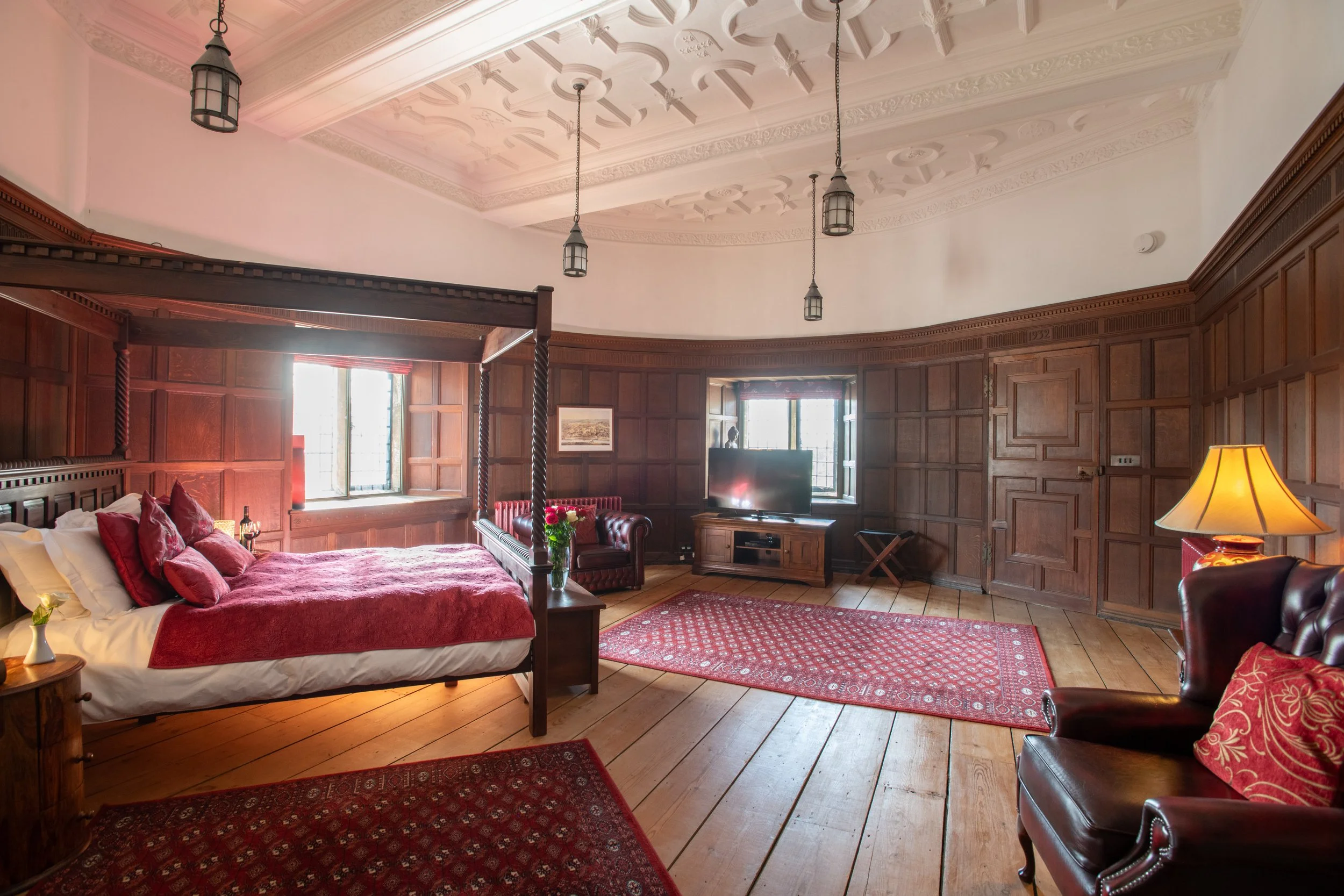 A luxurious bedroom with a high, ornately decorated ceiling and large windows. The room features wooden wall paneling, a four-poster bed with red bedding and pillows, a wooden dresser with a television, and vintage-style hanging lamps. There are red 