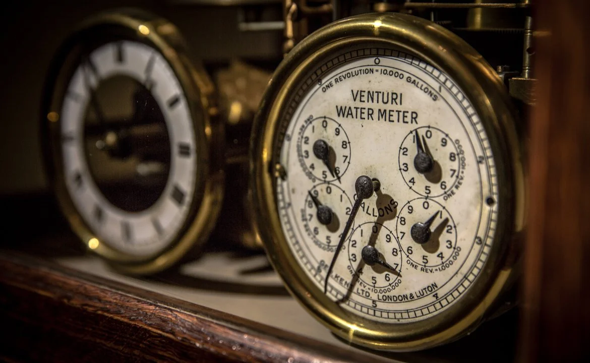 Close-up of vintage water meters with brass casing and white faces with black and red markings, installed on a wooden surface.