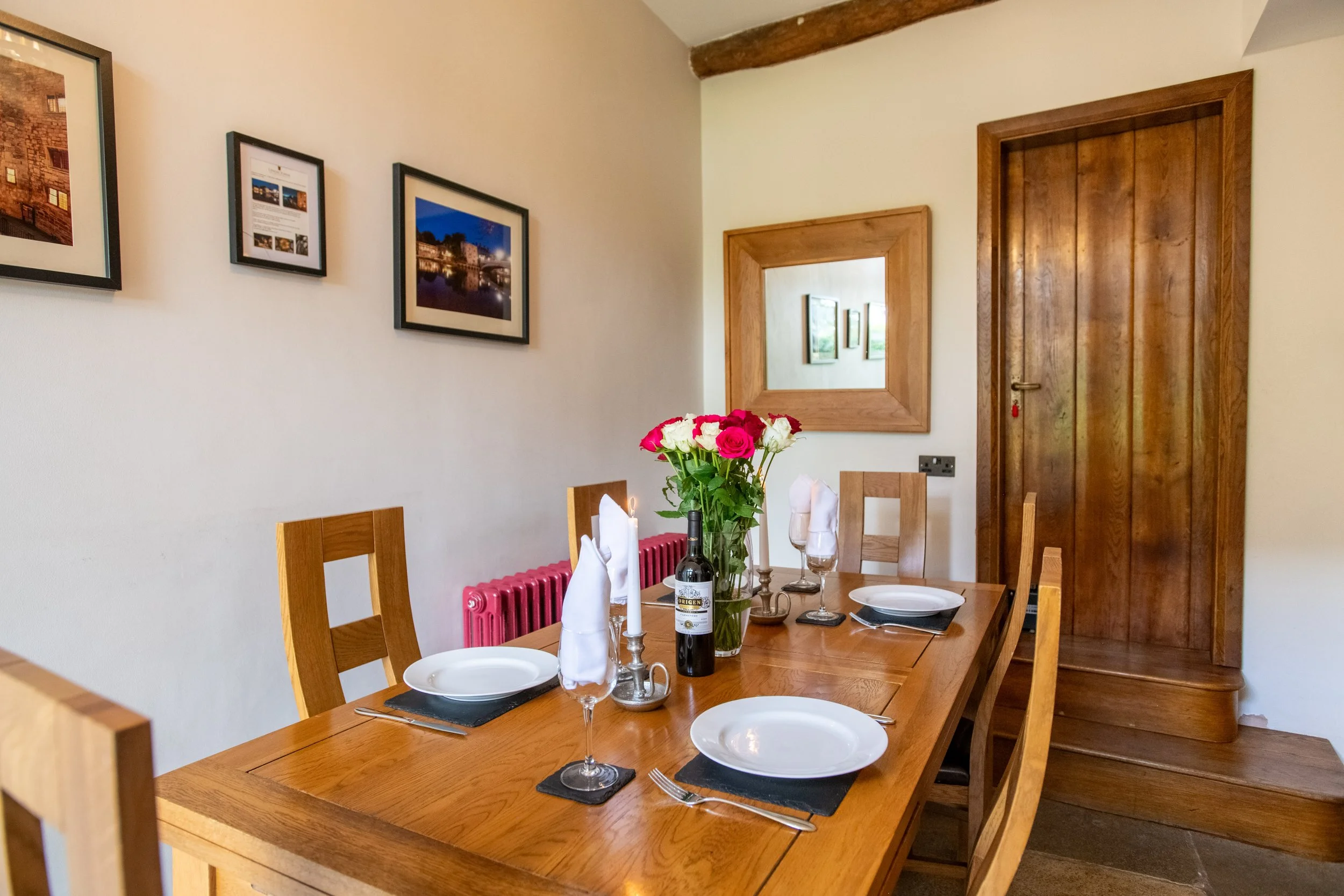 Dining room with a wooden table set for four, decorated with a vase of pink and white roses, wine bottles, and candlesticks. The walls feature framed pictures, a square mirror, and there is a wooden door and exposed ceiling beam.
