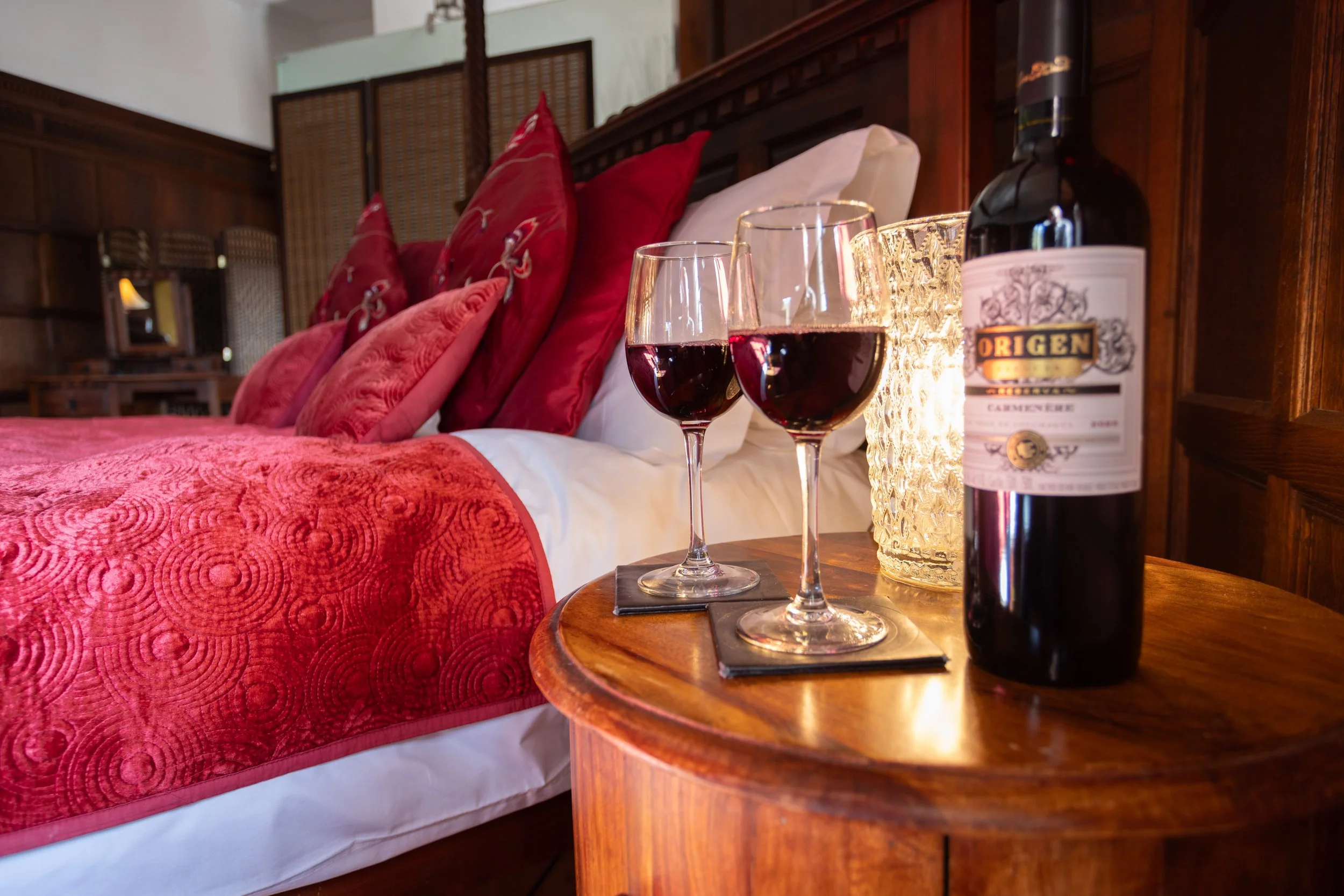 Two glasses of red wine on a wooden nightstand beside a bed with red and patterned pillows. A bottle of wine and a lit candle are also on the nightstand. The bed has a red quilt and white sheets, with a wooden headboard and wood-paneled walls.