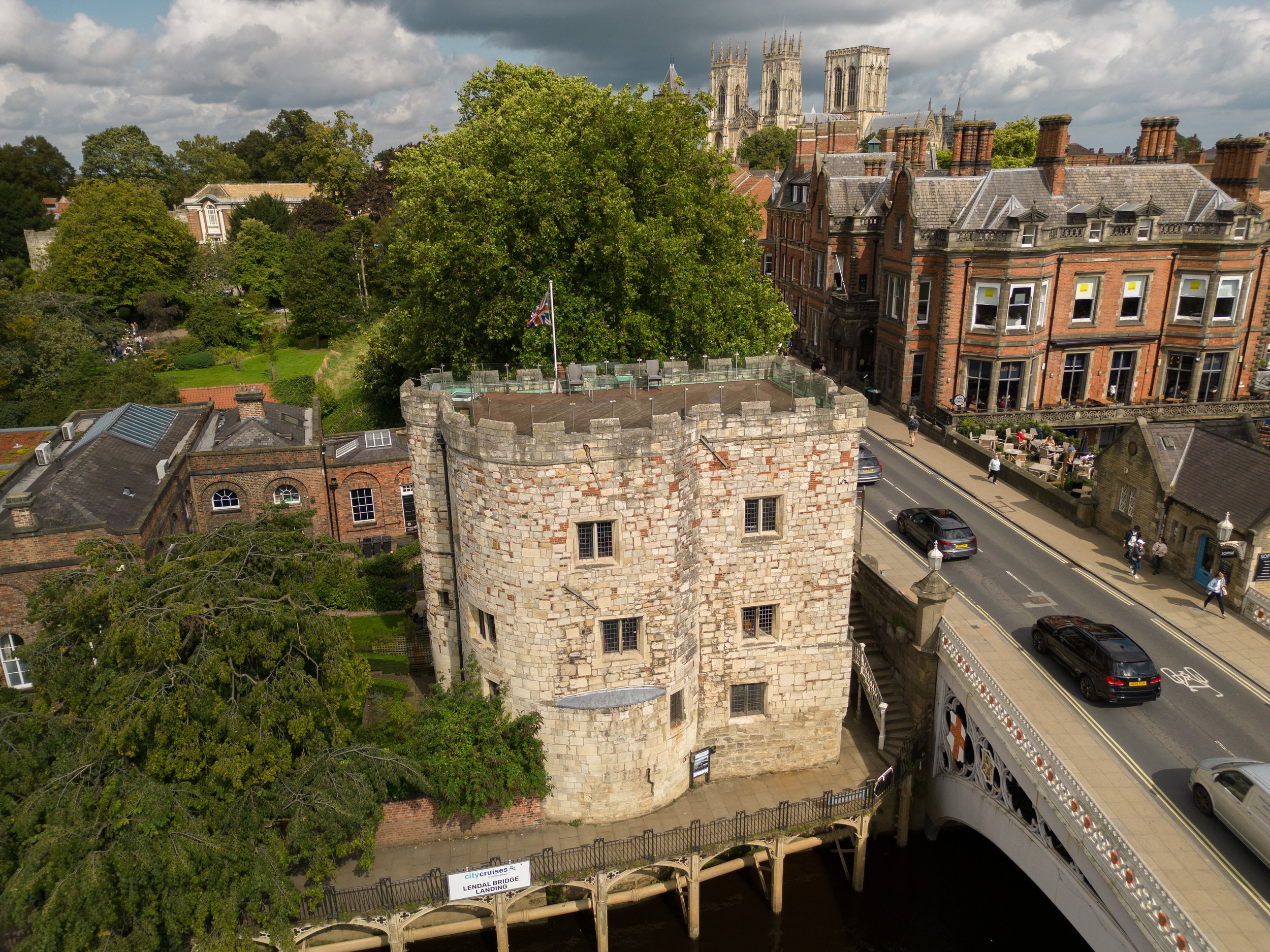 Aerial view of a historic stone tower with a flag, large trees, and red-brick buildings, with a city church in the background. A street runs alongside the buildings with cars and people walking, and a bridge labeled 'Lendal Bridge Landing' crosses a 
