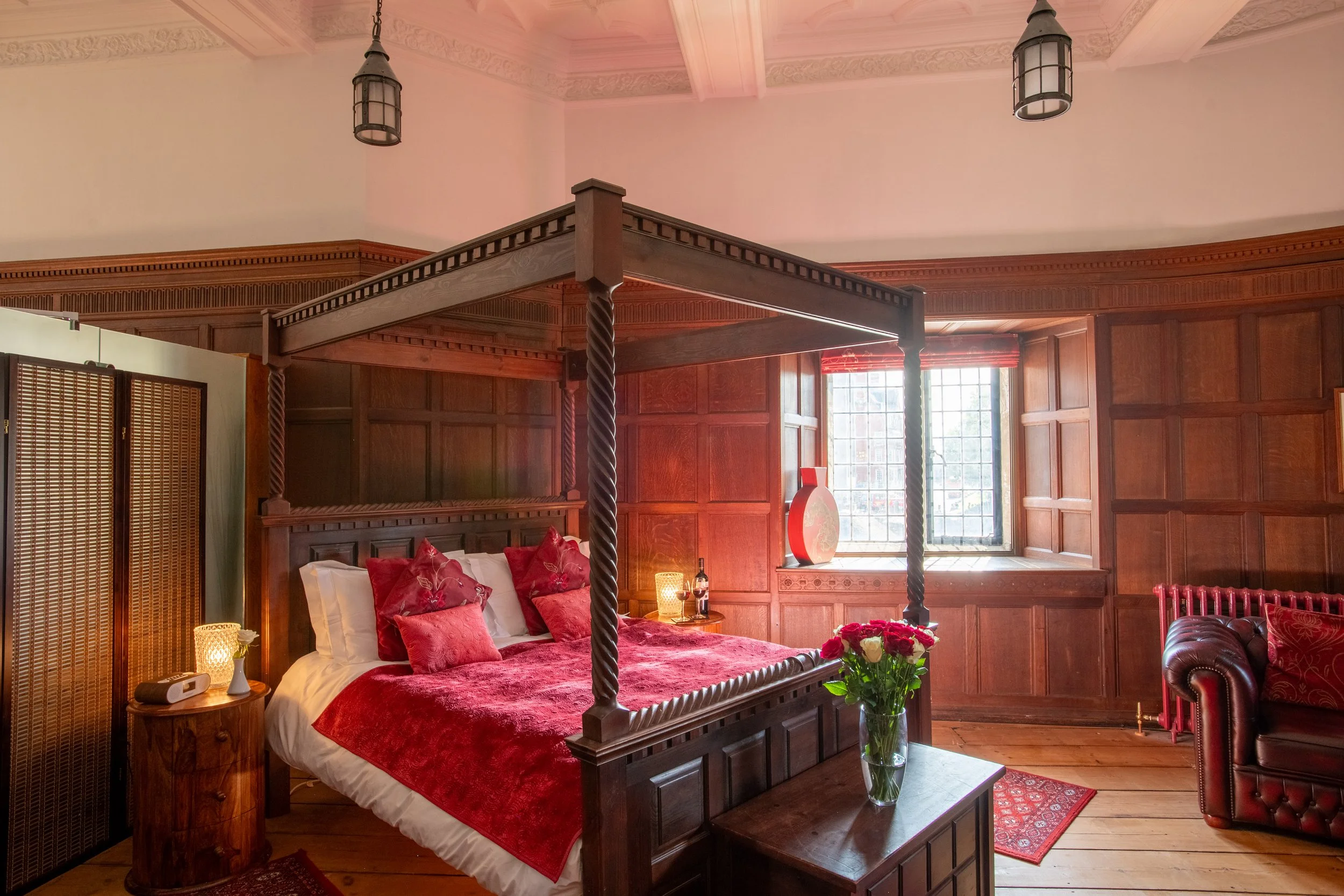 A bedroom with a four-poster bed with carved wood posts and red bedding, a nightstand with a lamp, a vase of flowers on a table, wood-paneled walls, large window with natural light, and ceiling lights.