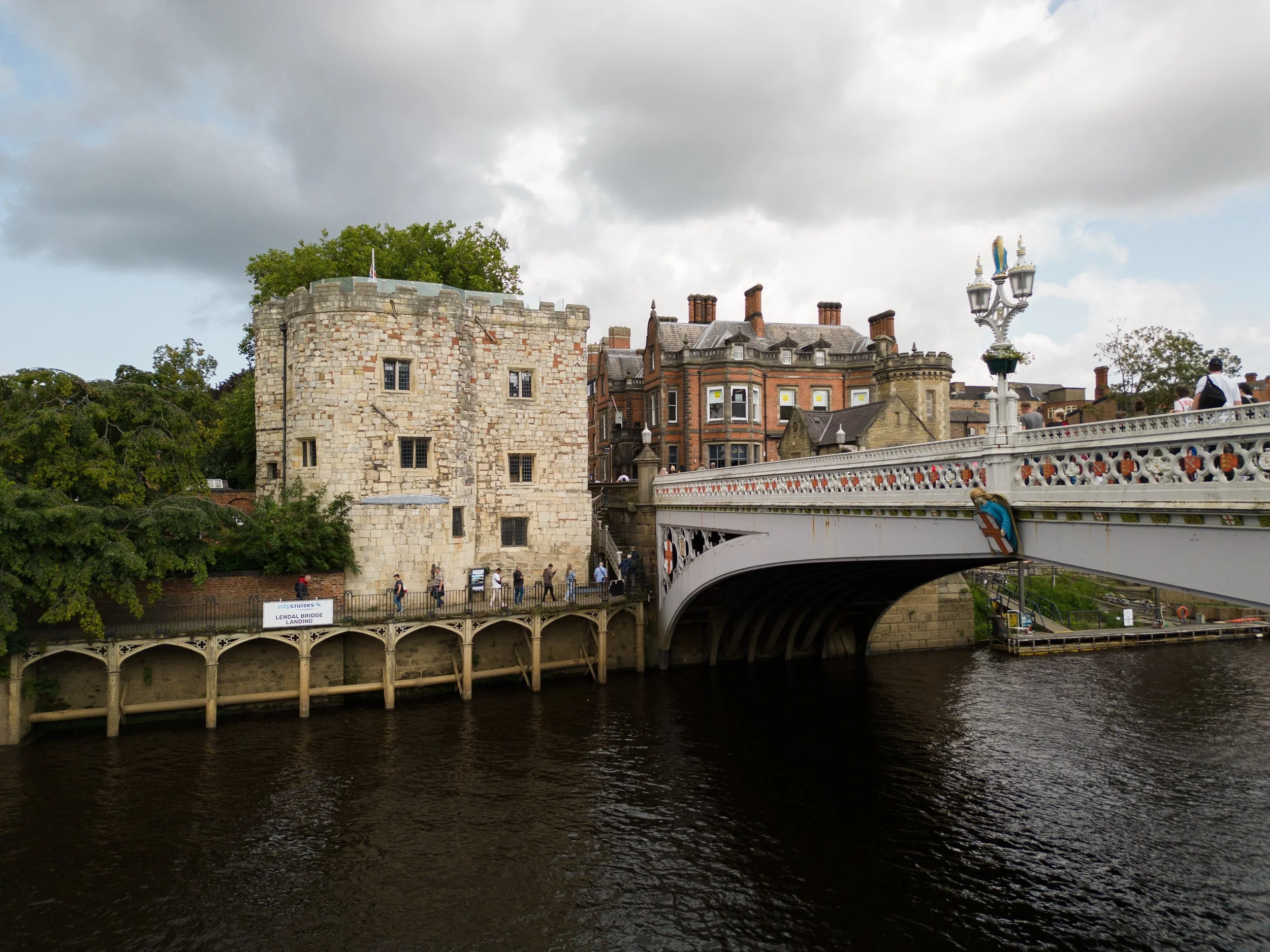 Lendal Bridge over a river with historic buildings and a stone tower in York, England, cloudy sky, group of people walking on the bridge and along the riverbank.