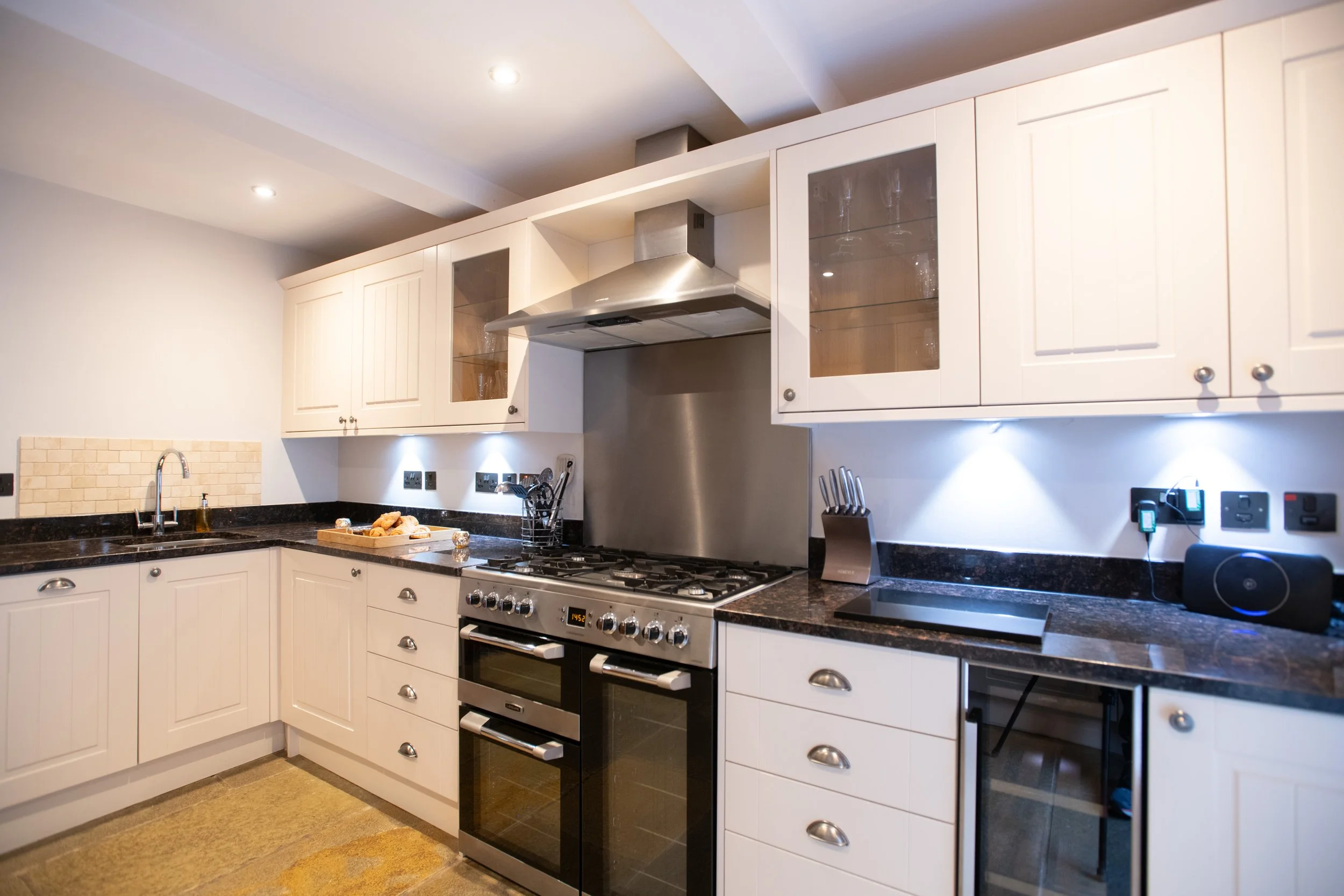 A modern kitchen with white cabinets, a black granite countertop, and stainless steel appliances including a stove and range hood. There is under-cabinet lighting, a sink with a curved faucet, and various kitchen utensils on the counter. The ceiling 