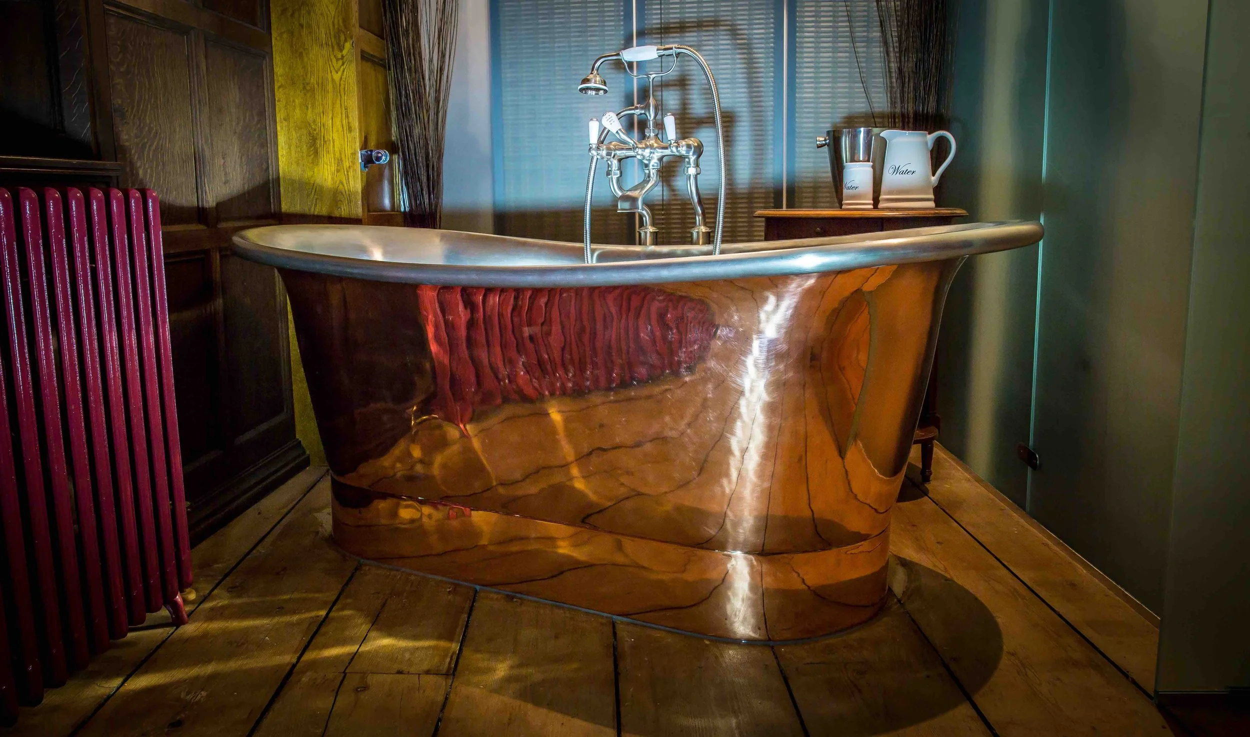 A vintage-style clawfoot bathtub with a wooden grain exterior, situated in a room with dark wood paneling, a green-painted wall, and a window with blinds. To the right, there's a small table with a pitcher and a glass labeled 'Water'.