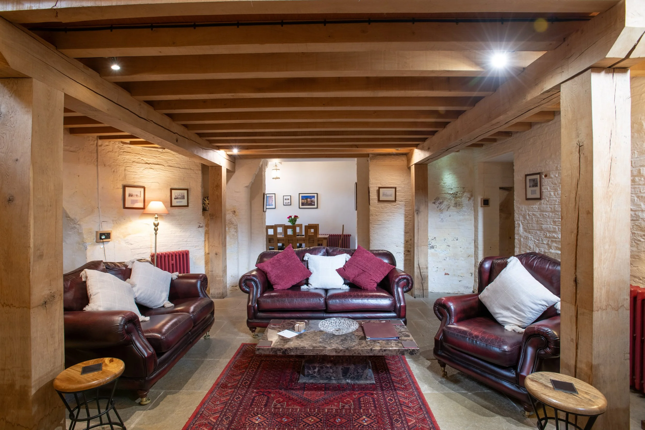 Living room with leather sofas, white and red pillows, wooden beams on ceiling, framed pictures on stone wall, table with books, small side tables, rug, and a lamp.