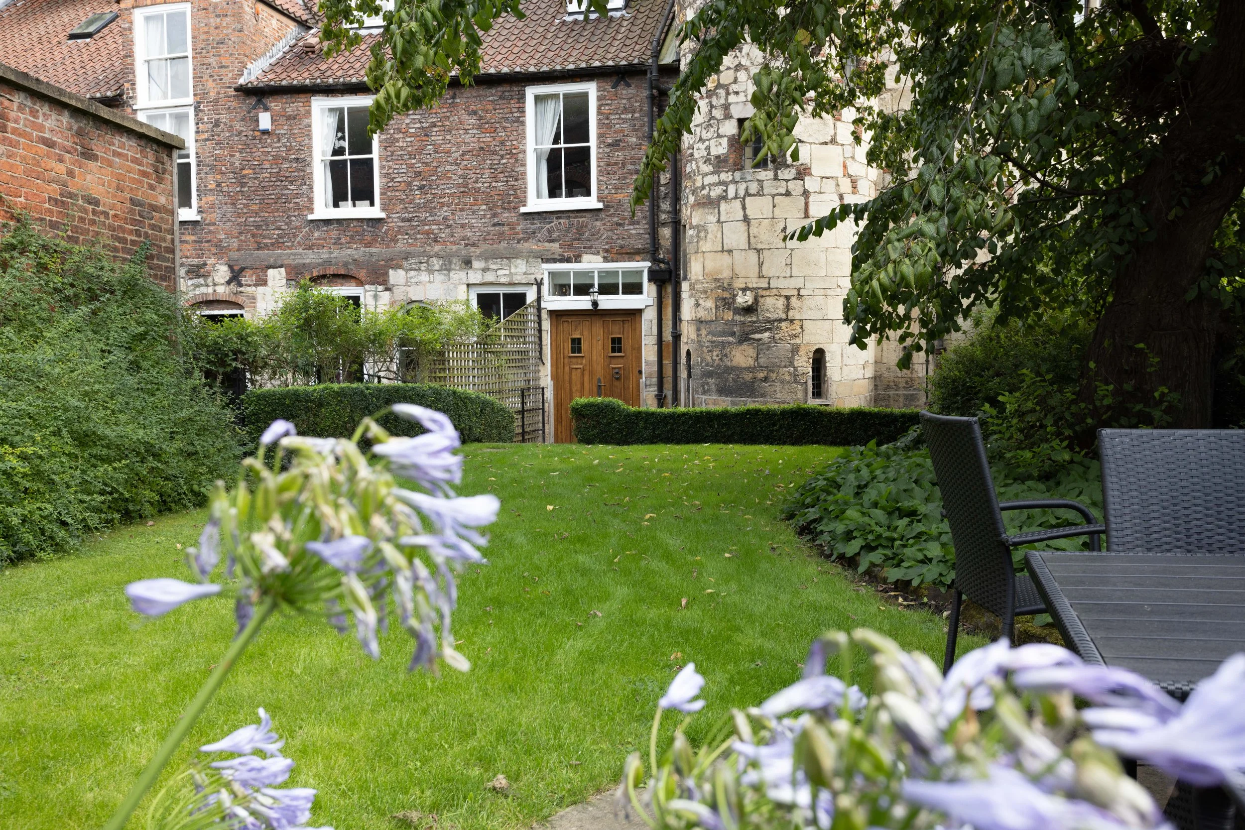 A garden with green grass, purple flowers in the foreground, and a brick and stone house with tall white-framed windows and a wooden door in the background. There are trees and bushes around the garden.