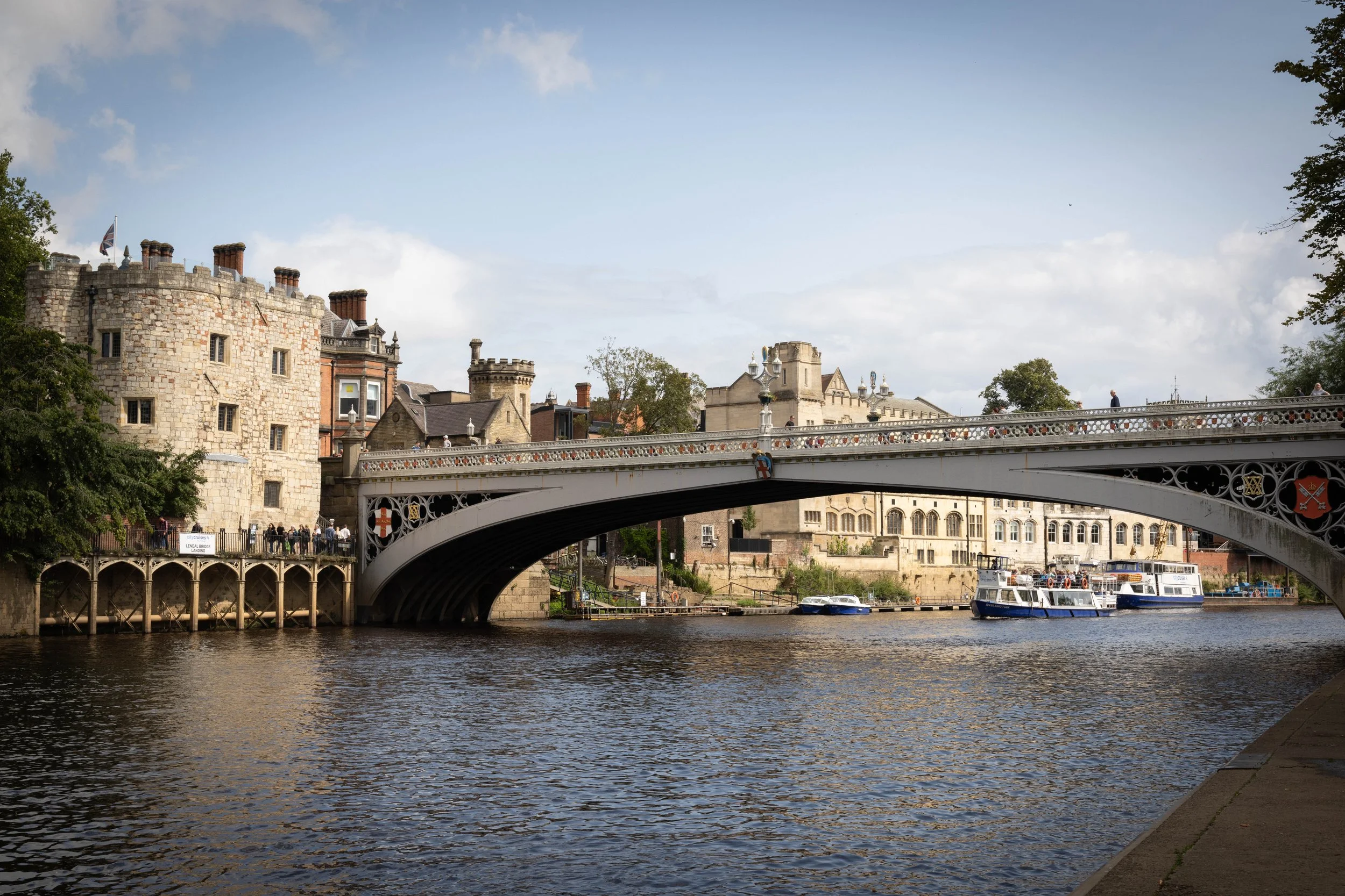 View of the River Thames showing a bridge with people walking, old buildings with chimneys in the background, and a boat on the river.