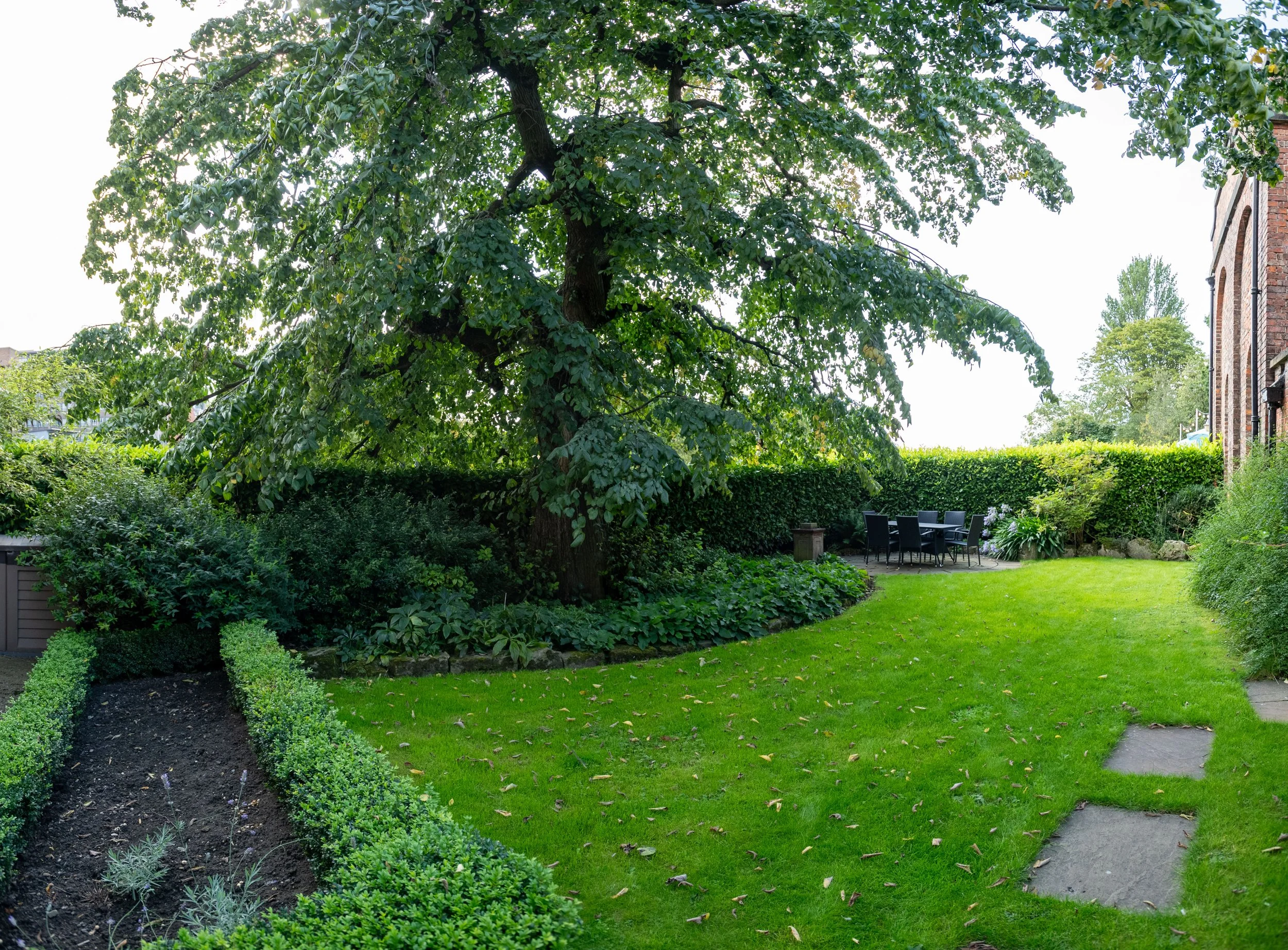 A lush green backyard garden with a large leafy tree, manicured bushes, a circular patio with outdoor dining furniture, and a brick building on the right.