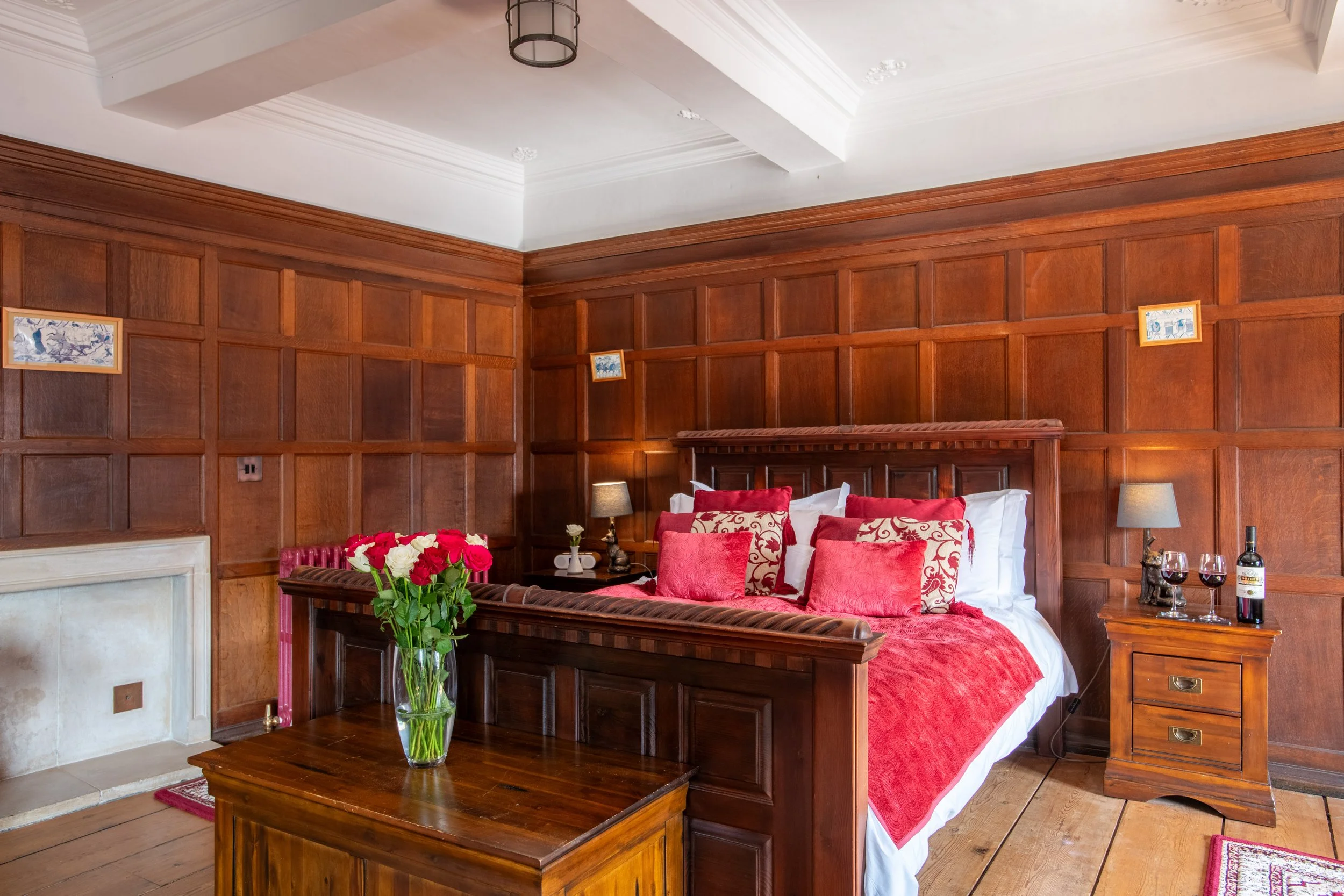 A bedroom with wooden paneled walls, a bed with red and white bedding, a bouquet of red and white flowers on a wooden coffee table, and a pair of wine glasses with a bottle. There are two lamps on nightstands and small framed pictures on the wall.