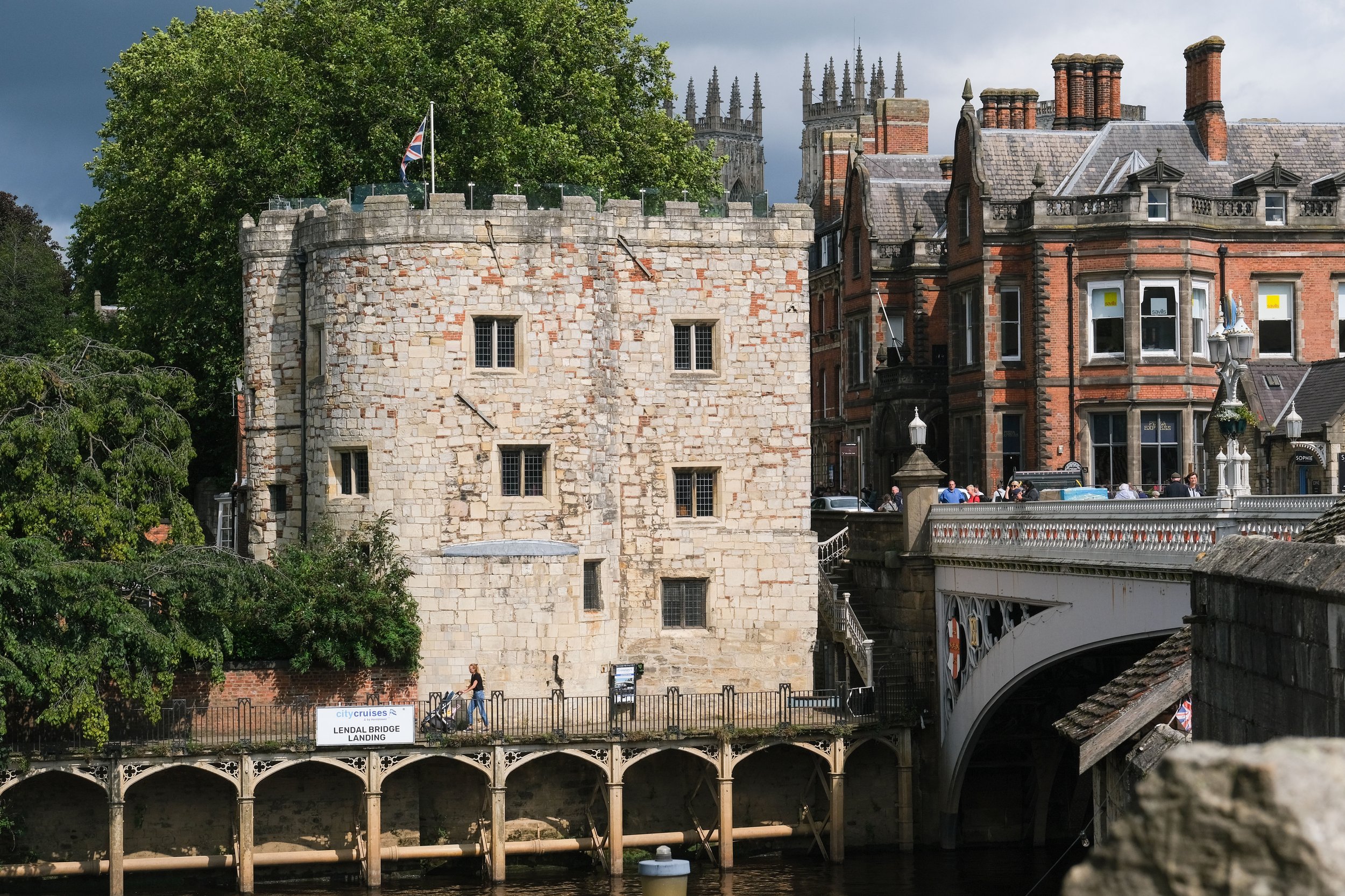 Historical buildings with a stone tower, brick and stone architecture, a bridge with decorative arches, and a sign reading 'Lendal Bridge Landing' amid green trees and a cloudy sky.
