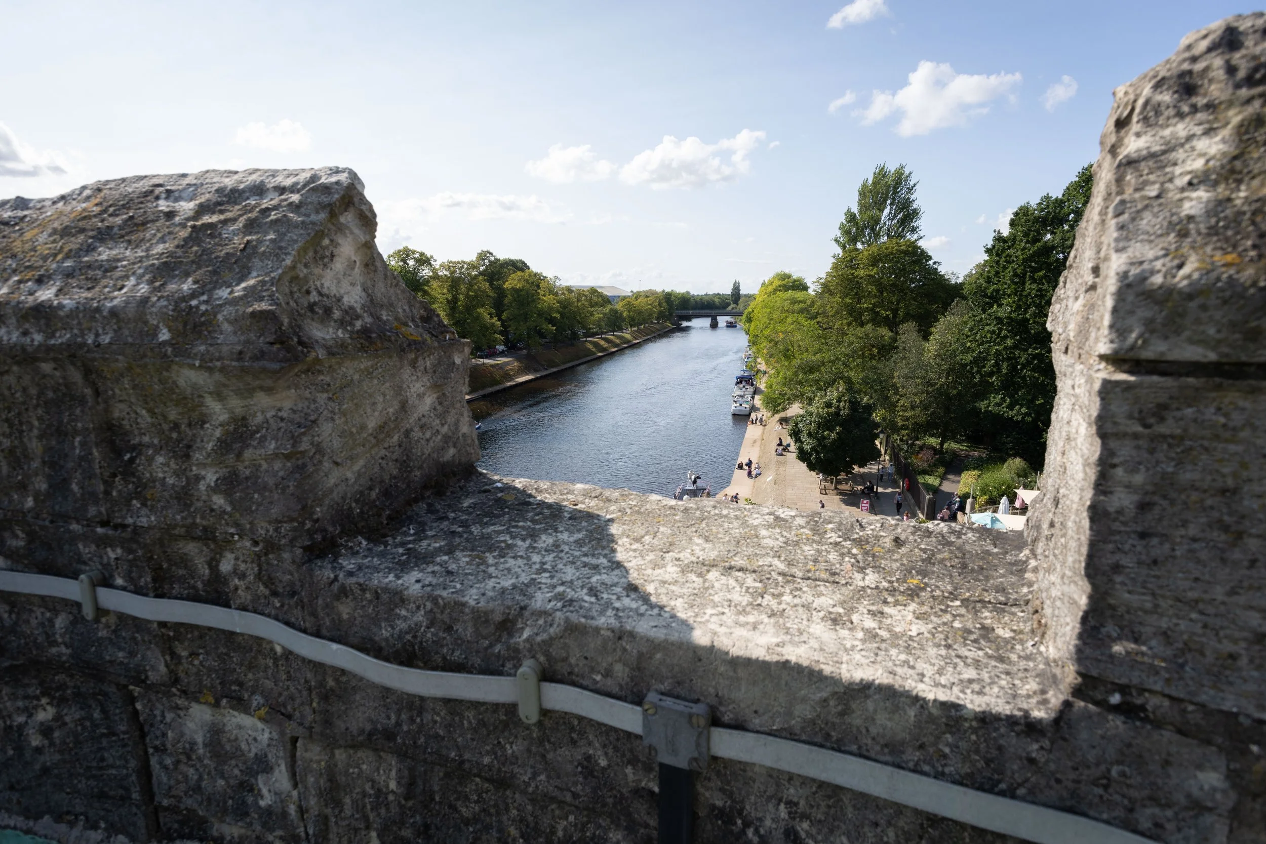 View through a gap in a stone wall looking at a river with boats, green trees, and a bridge in the distance on a sunny day.