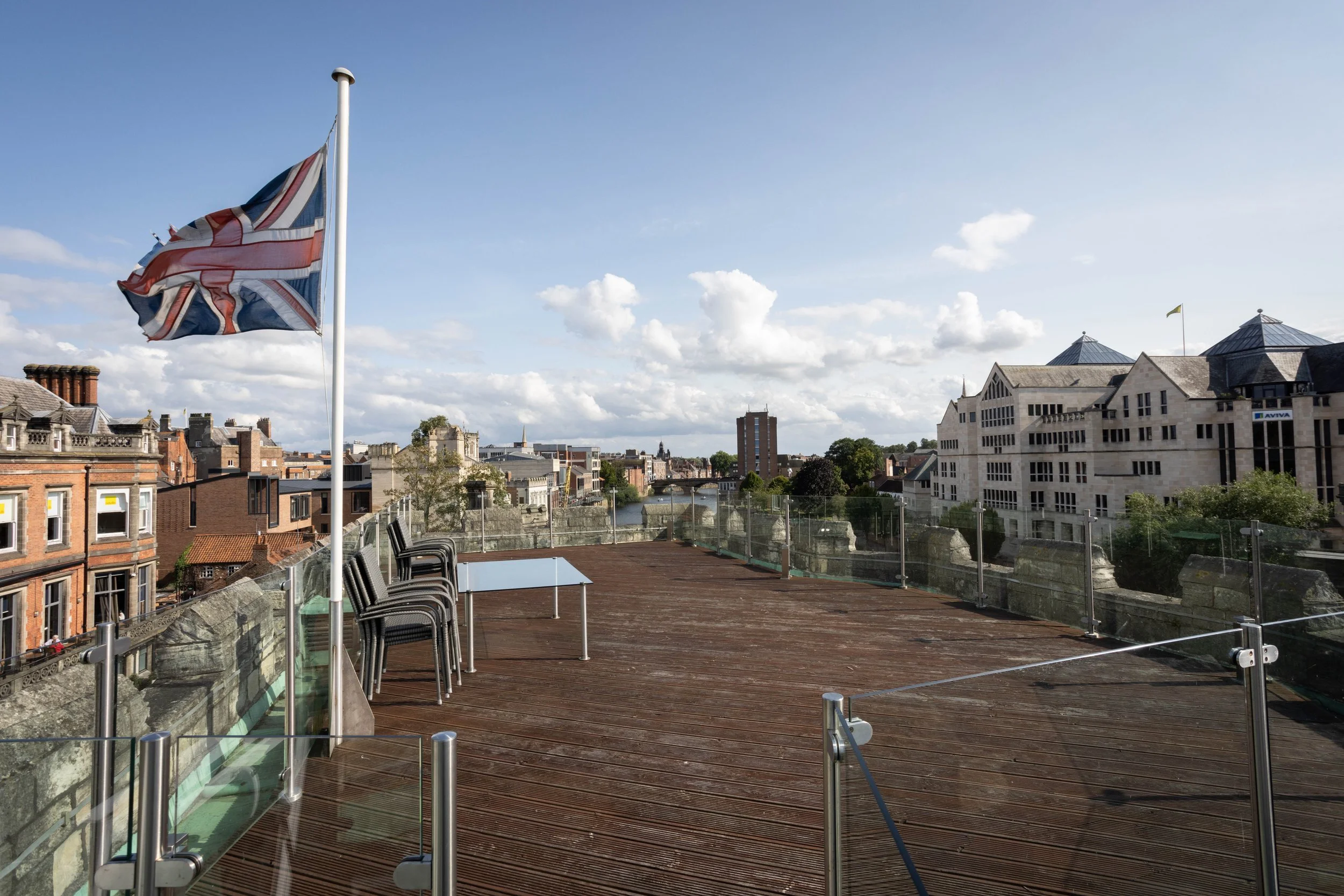 A rooftop viewpoint showing a Union Jack flag waving, with a cityscape in the background including residential buildings, a river, and a clear blue sky with some clouds.