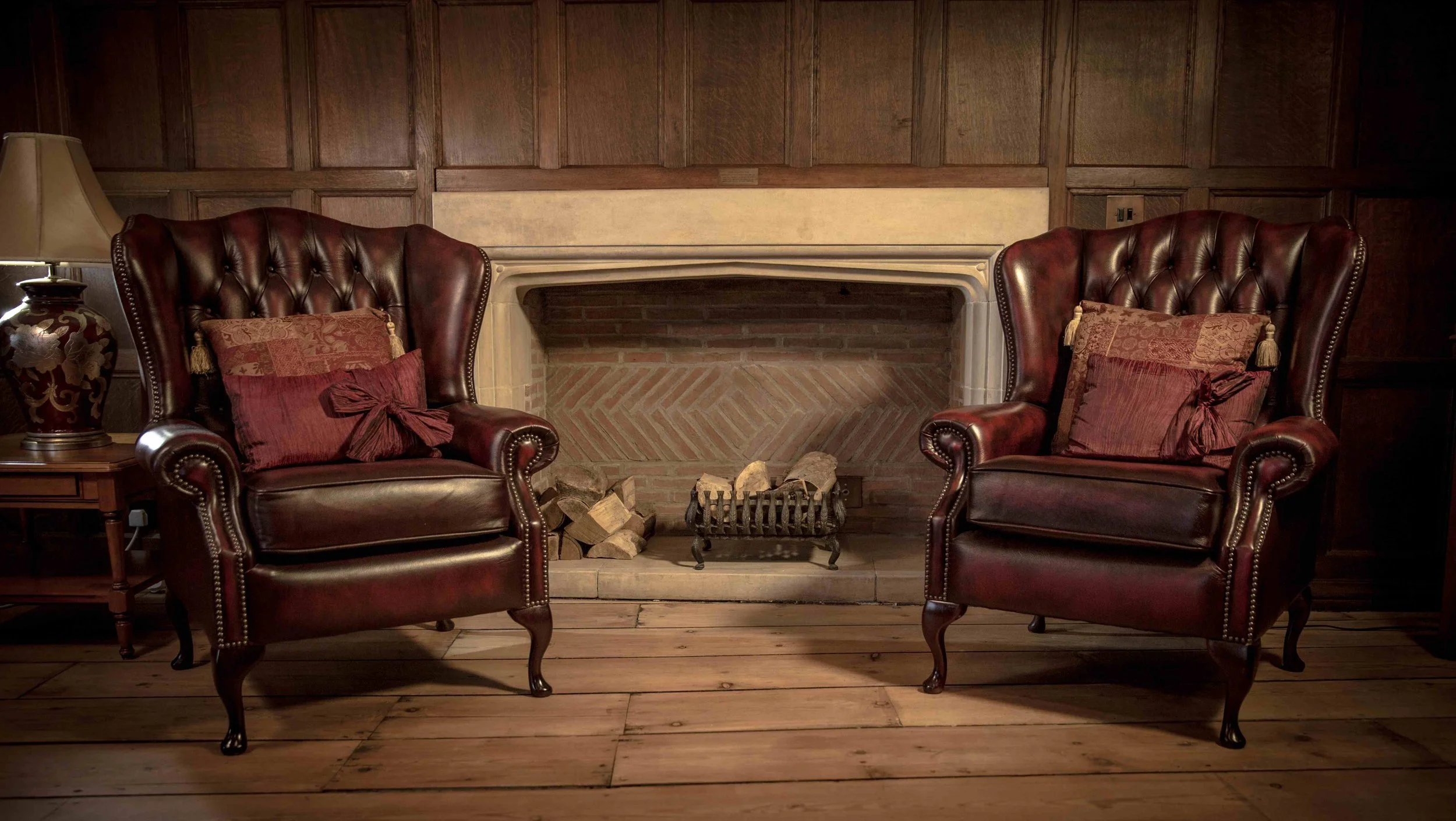 A cozy living room with two leather armchairs with red pillows, a fireplace, a wooden floor, a side table with a lamp, and wood-paneled walls.