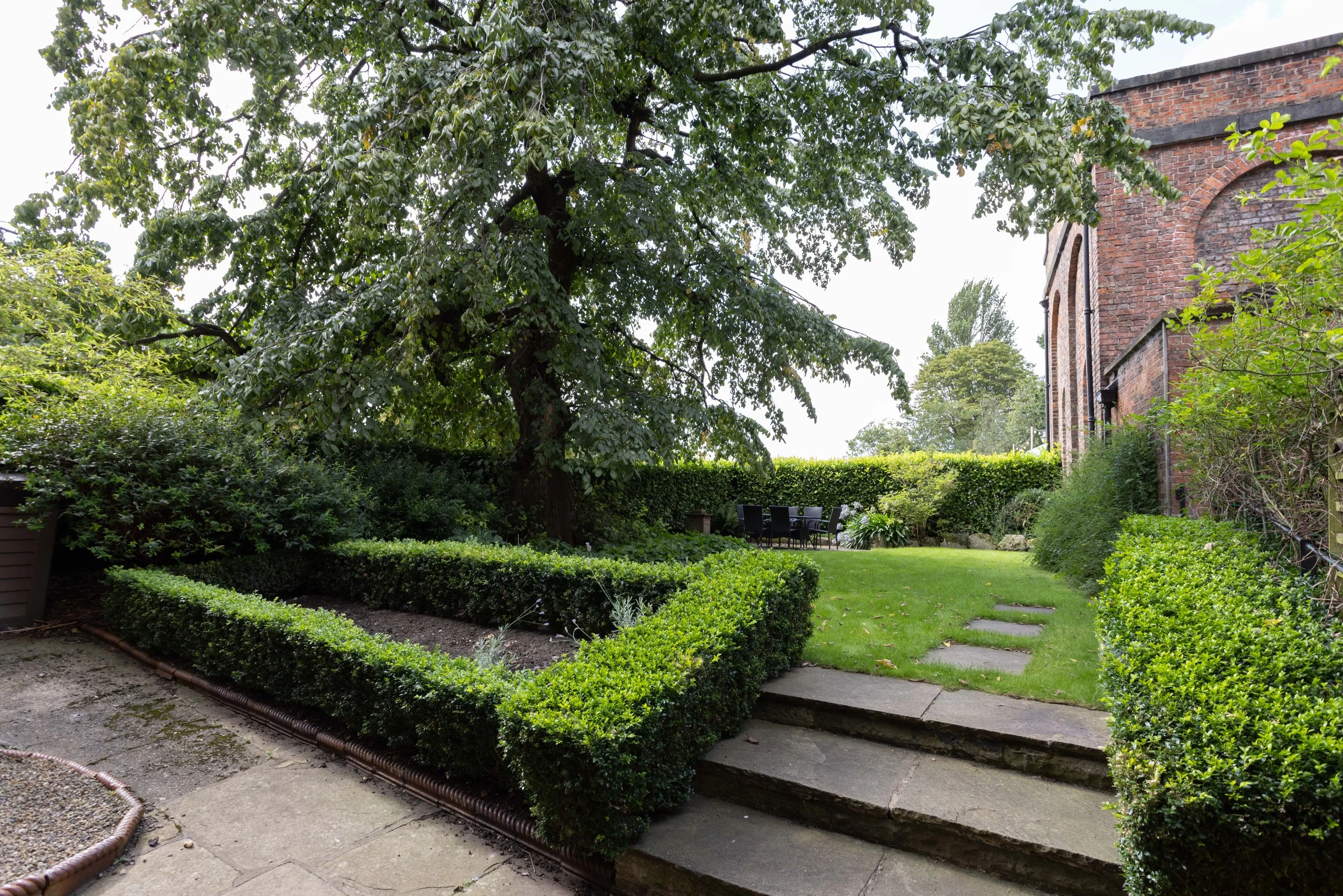 Green garden with trimmed bushes and a large tree, brick building on the right, outdoor table and chairs, concrete steps, and a treeline in the background.