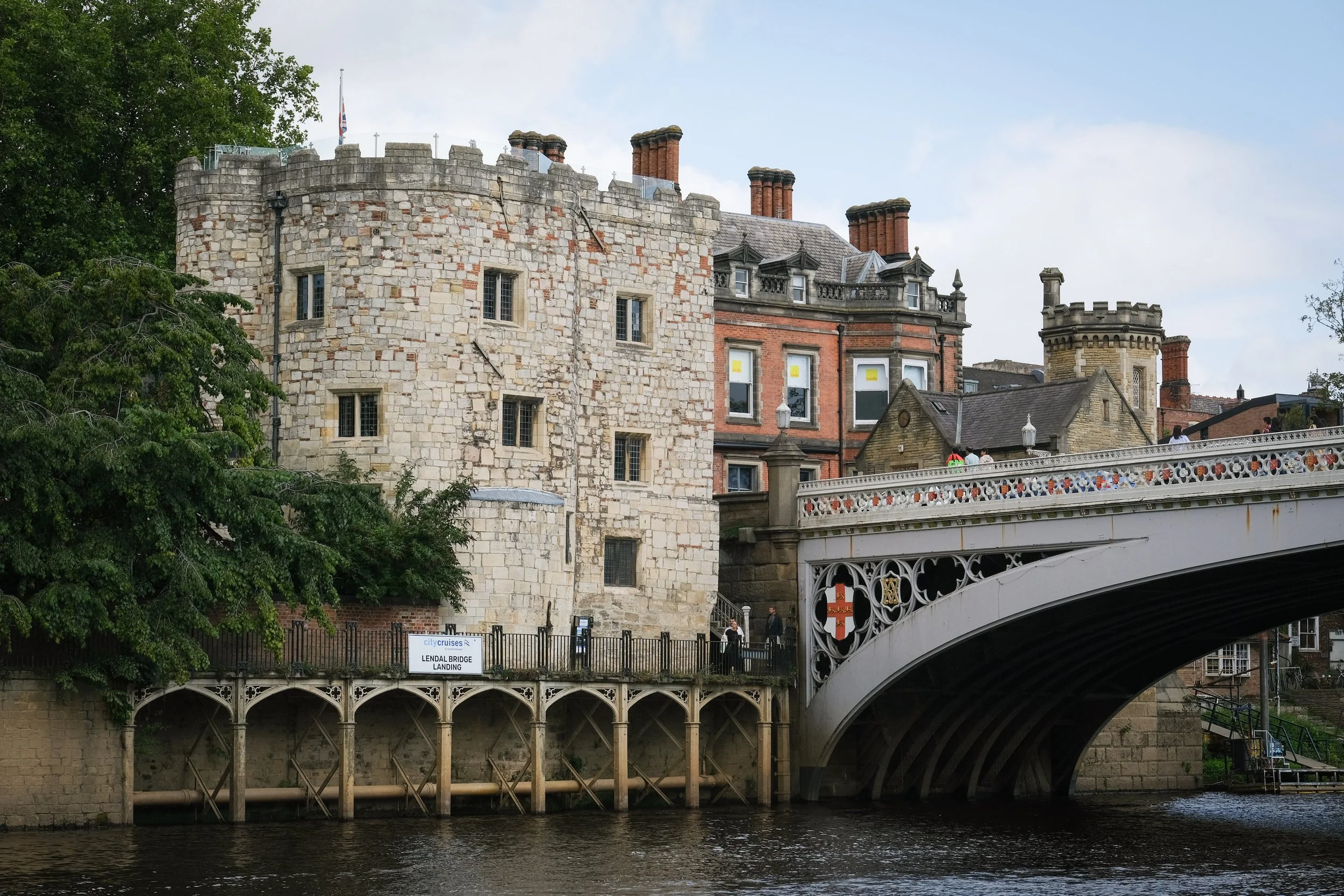 Historic stone castle building with multiple chimneys and ornate details, located near a river with a decorative bridge in the foreground and green trees on one side.