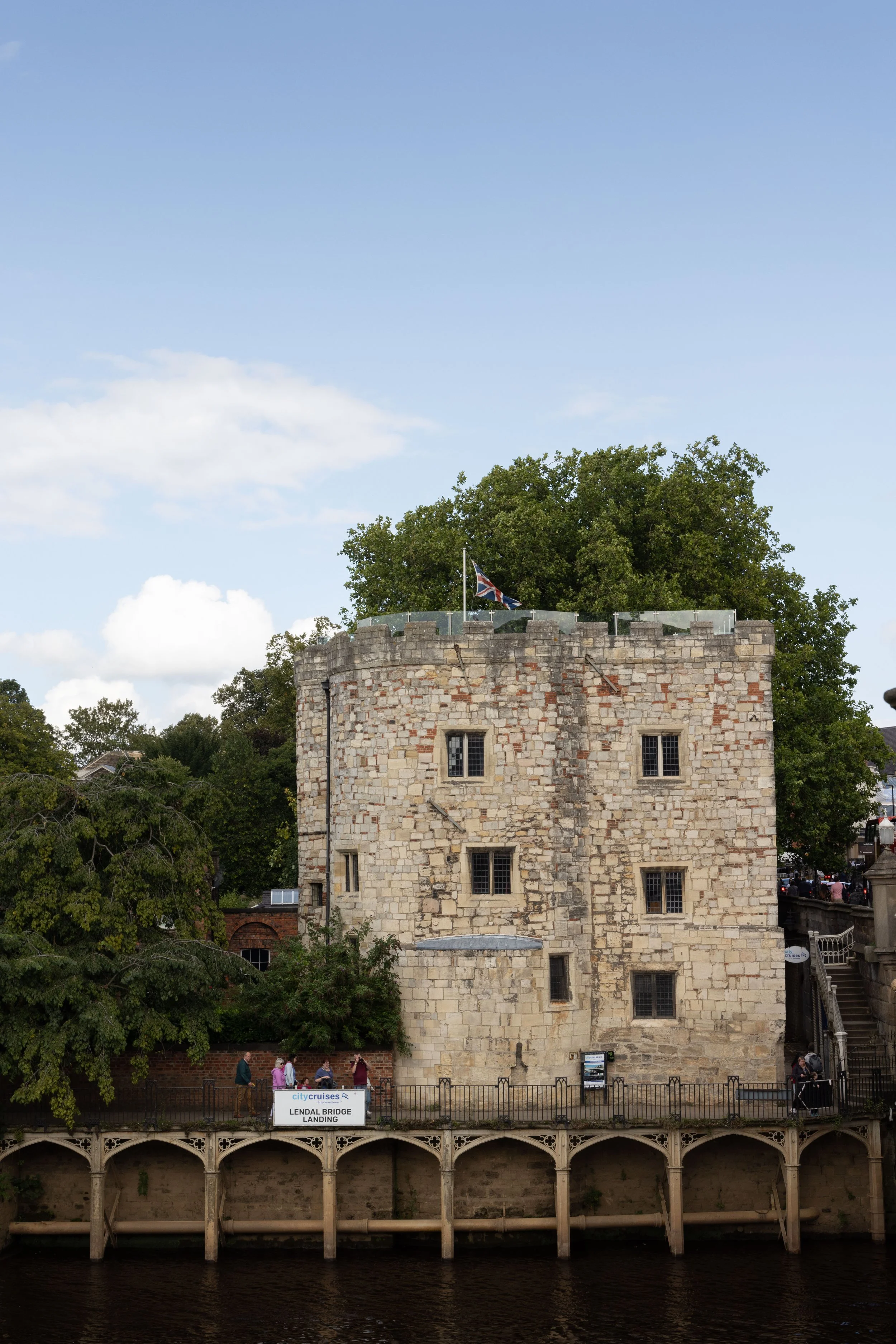 View of Landal Bridge Landing with a stone tower, people walking along the walkway, and a sign indicating 'Lendal Bridge Landing' near the river, with trees and a partly cloudy sky in the background.