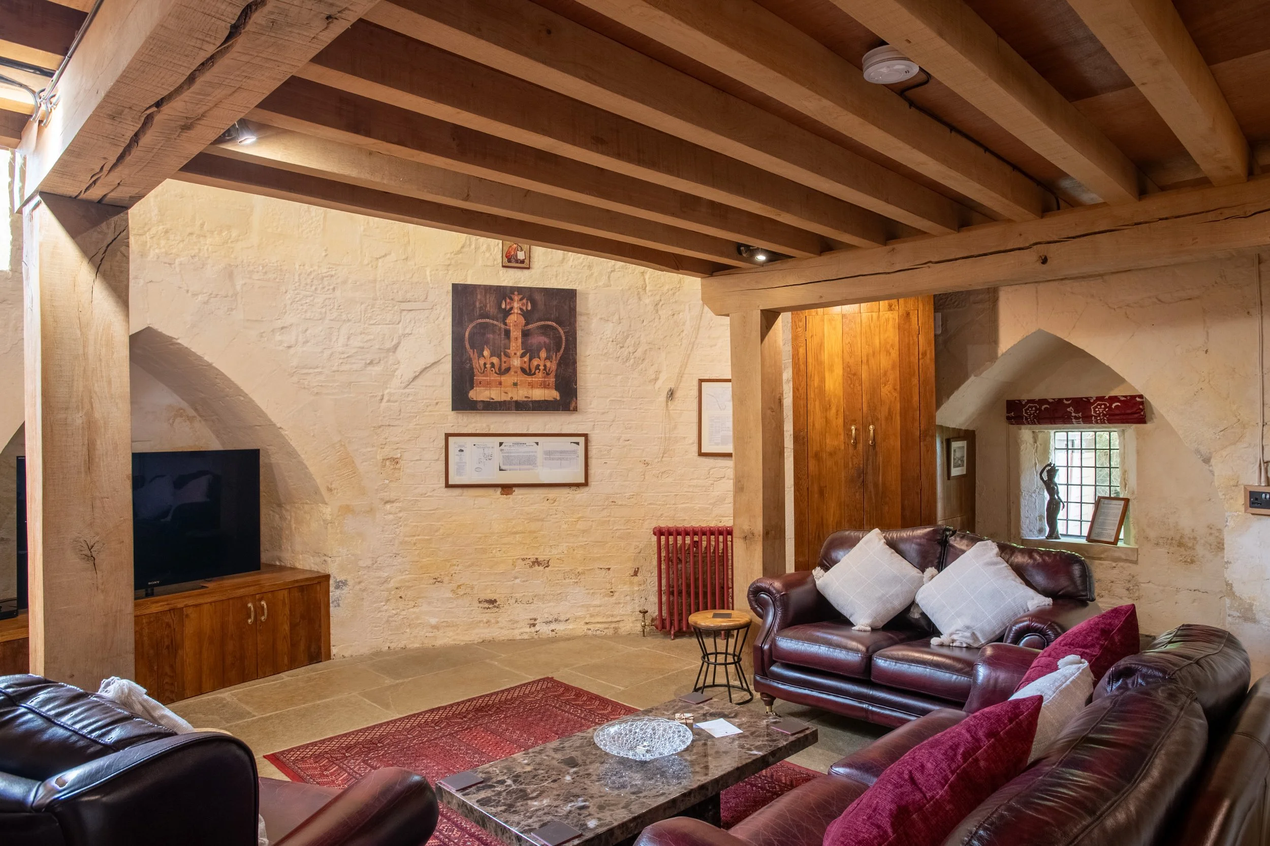 A cozy living room with exposed wooden beams on the ceiling, a leather sofa with white and patterned pillows, a marble coffee table, a red chair, and framed art on the brick wall.