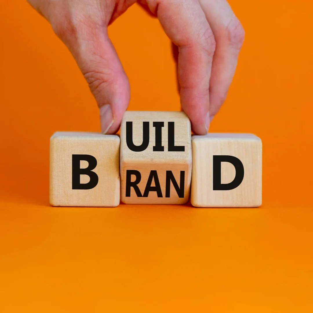 Wooden blocks spelling out 'BUILD YOUR BRAND' against an orange background, with a hand placing the middle block.