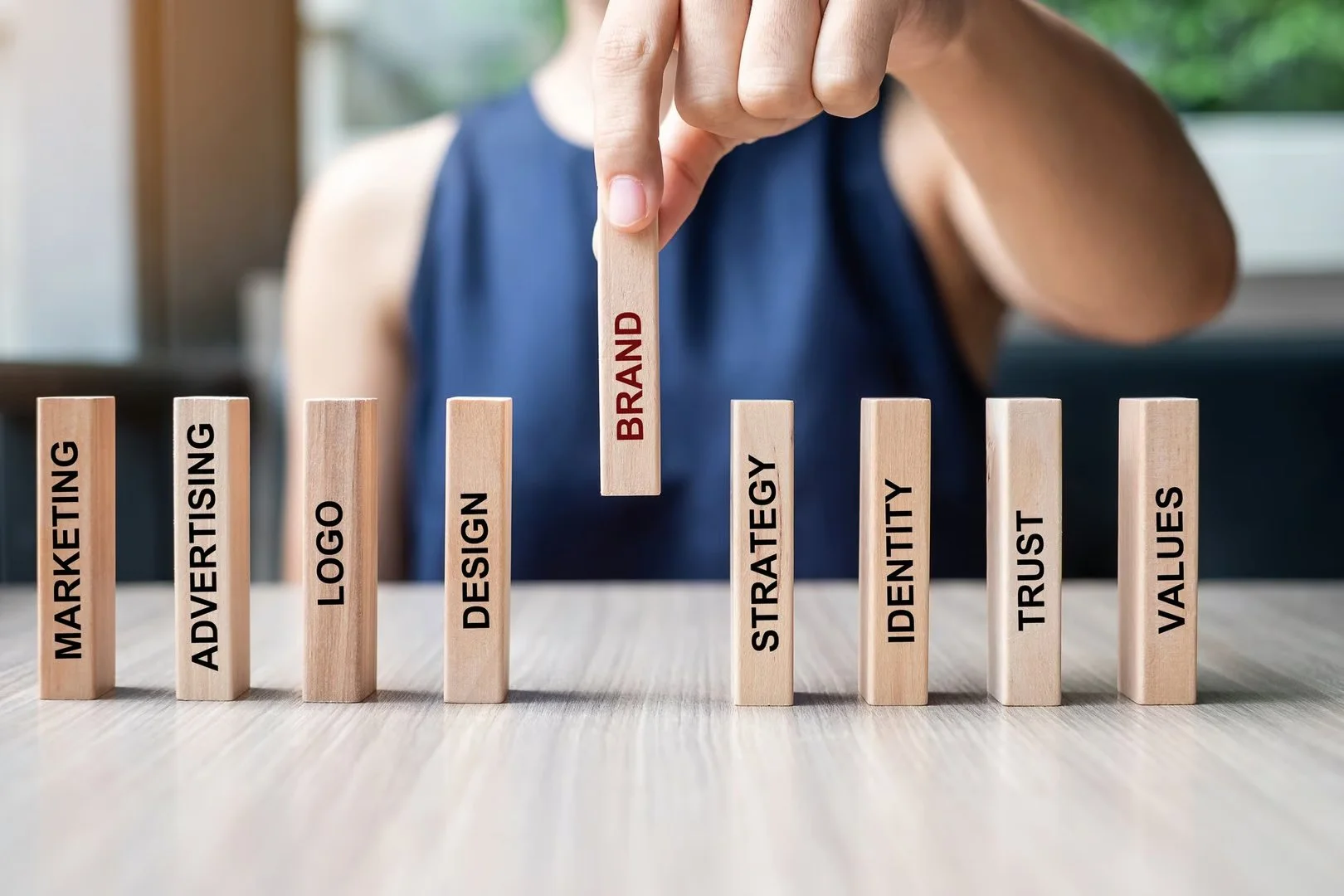 A person in a blue sleeveless shirt moves a wooden block labeled 'BRAND' among other blocks labeled 'MARKETING,' 'ADVERTISING,' 'LOGO,' 'DESIGN,' 'STRATEGY,' 'IDENTITY,' 'TRUST,' and 'VALUES' on a light wooden surface, with green foliage in the background.