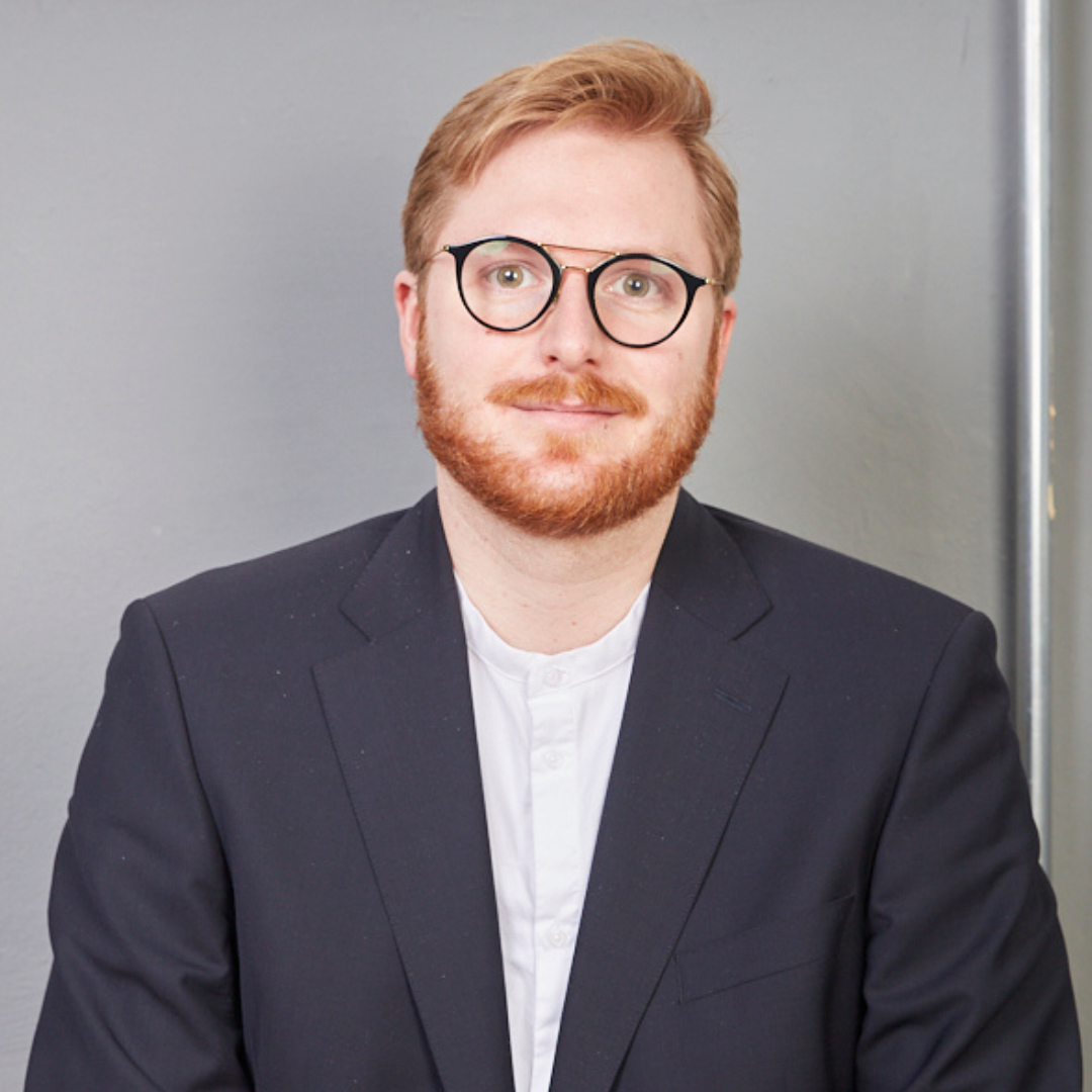 Ein Mann mit roten Haaren, Brille, Bart, schwarzer Jacke und weißen Hemd sitzt vor einer grauen Wand.