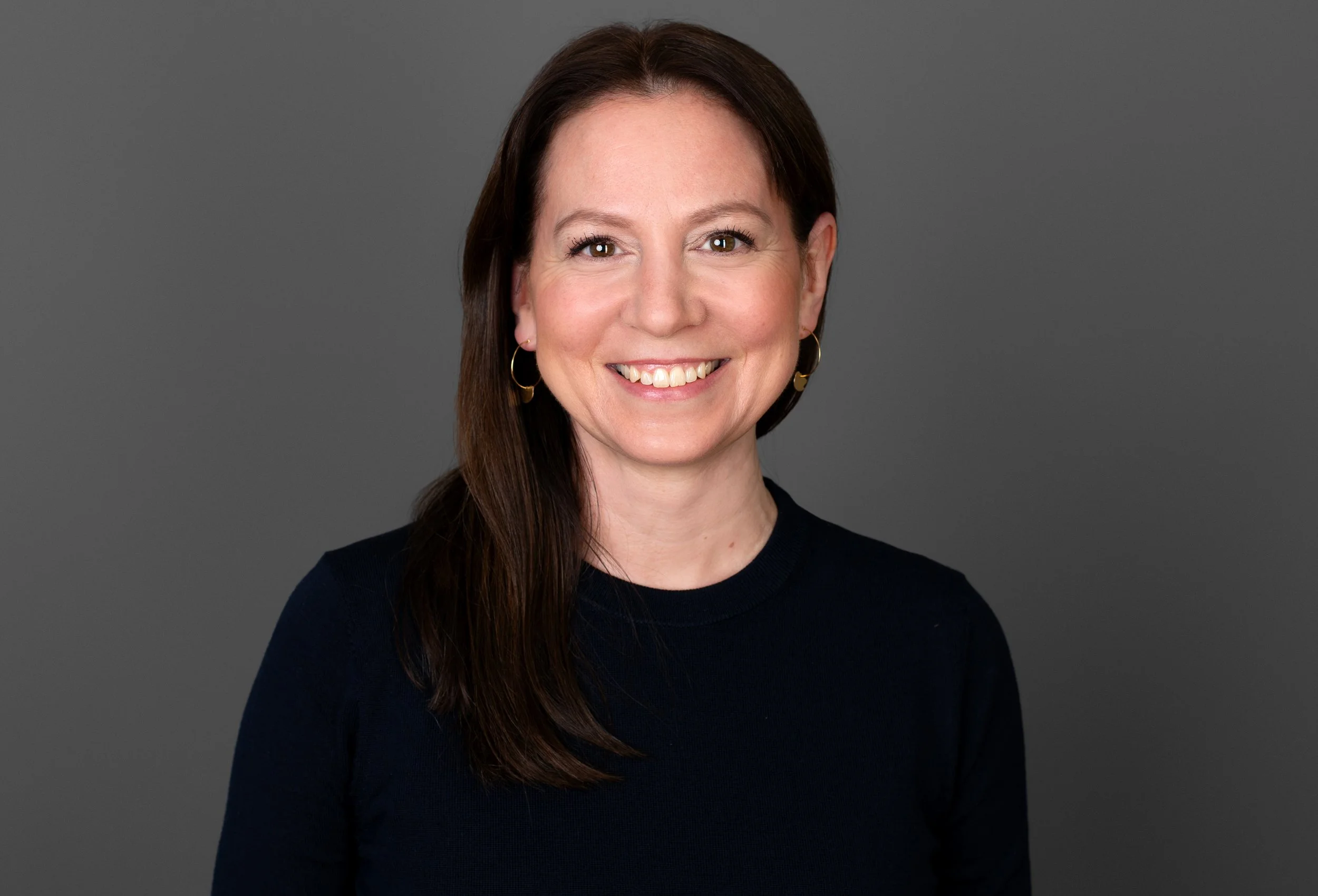 Eine lächelnde Frau mit braunem Haar vor grauem Hintergrund, trägt einen schwarzen Pullover und goldene Ohrringe.