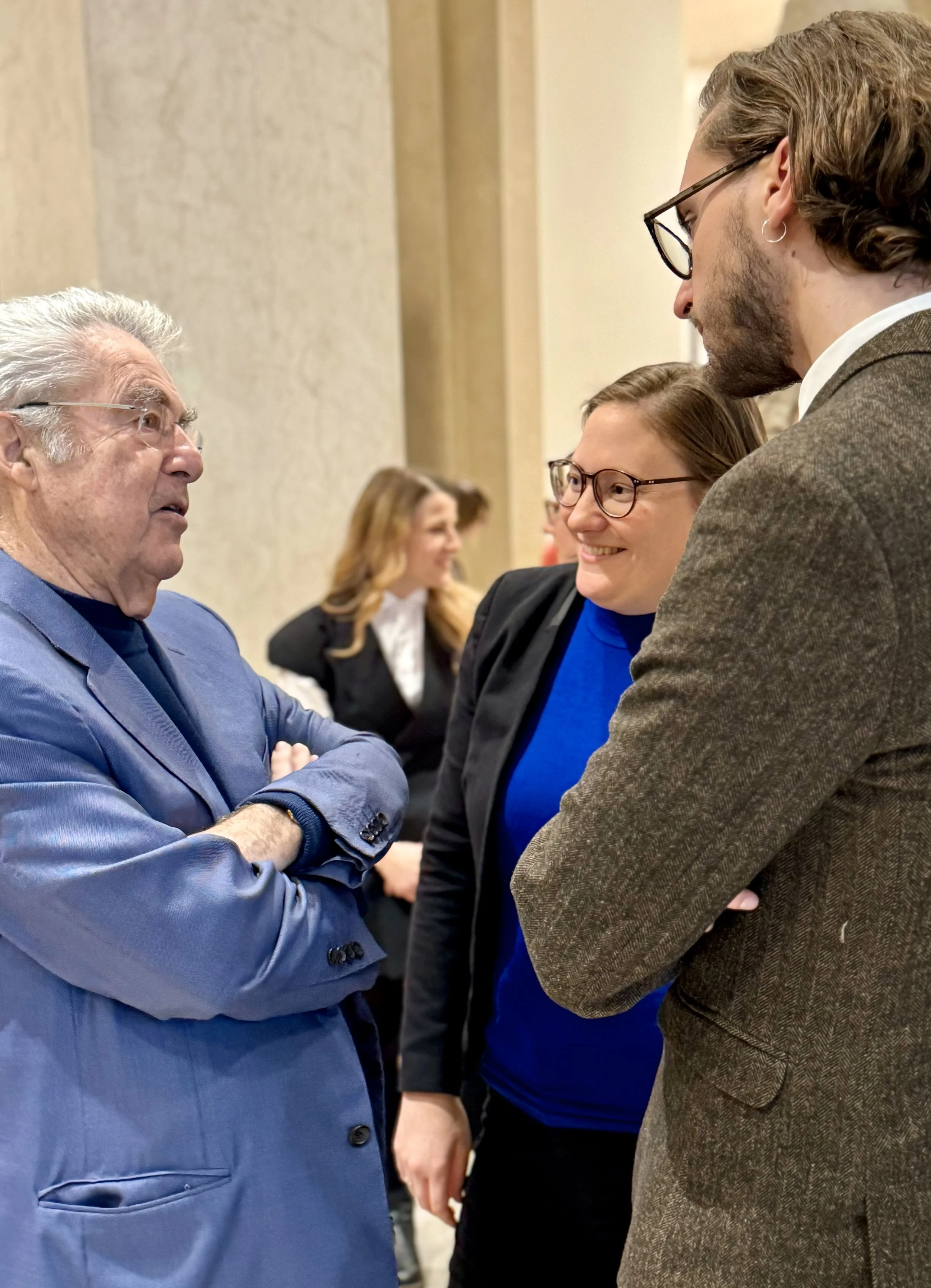 Drei Personen im Gespräch, alle tragen Brillen und sind in formeller Kleidung, in einem Raum mit anderen Menschen im Hintergrund.