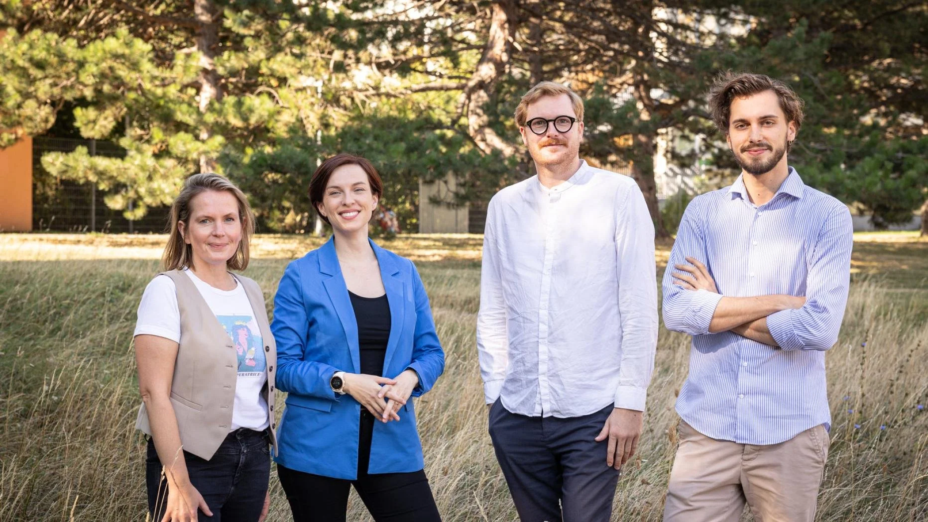 Fünf Menschen stehen im Freien auf einem Feld mit Bäumen im Hintergrund, lächeln und schauen in die Kamera.