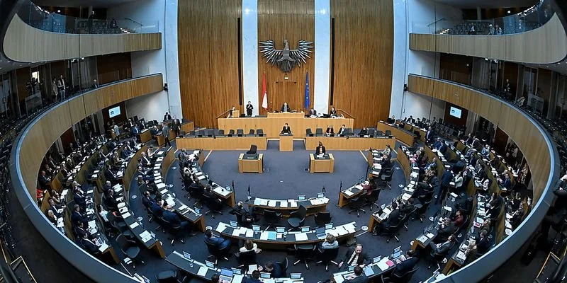 Der Deutsche Bundestag während einer Sitzung im Plenarsaal, mit Stühlen, Tischen und Abgeordneten, Bundesadler und Deutschland-Flagge im Hintergrund.