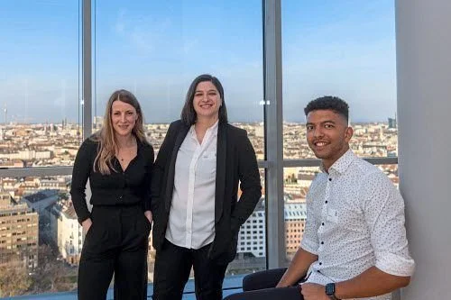 Drei Menschen in Businesskleidung stehen vor großen Fenstern mit Blick auf eine Stadt, lachen.