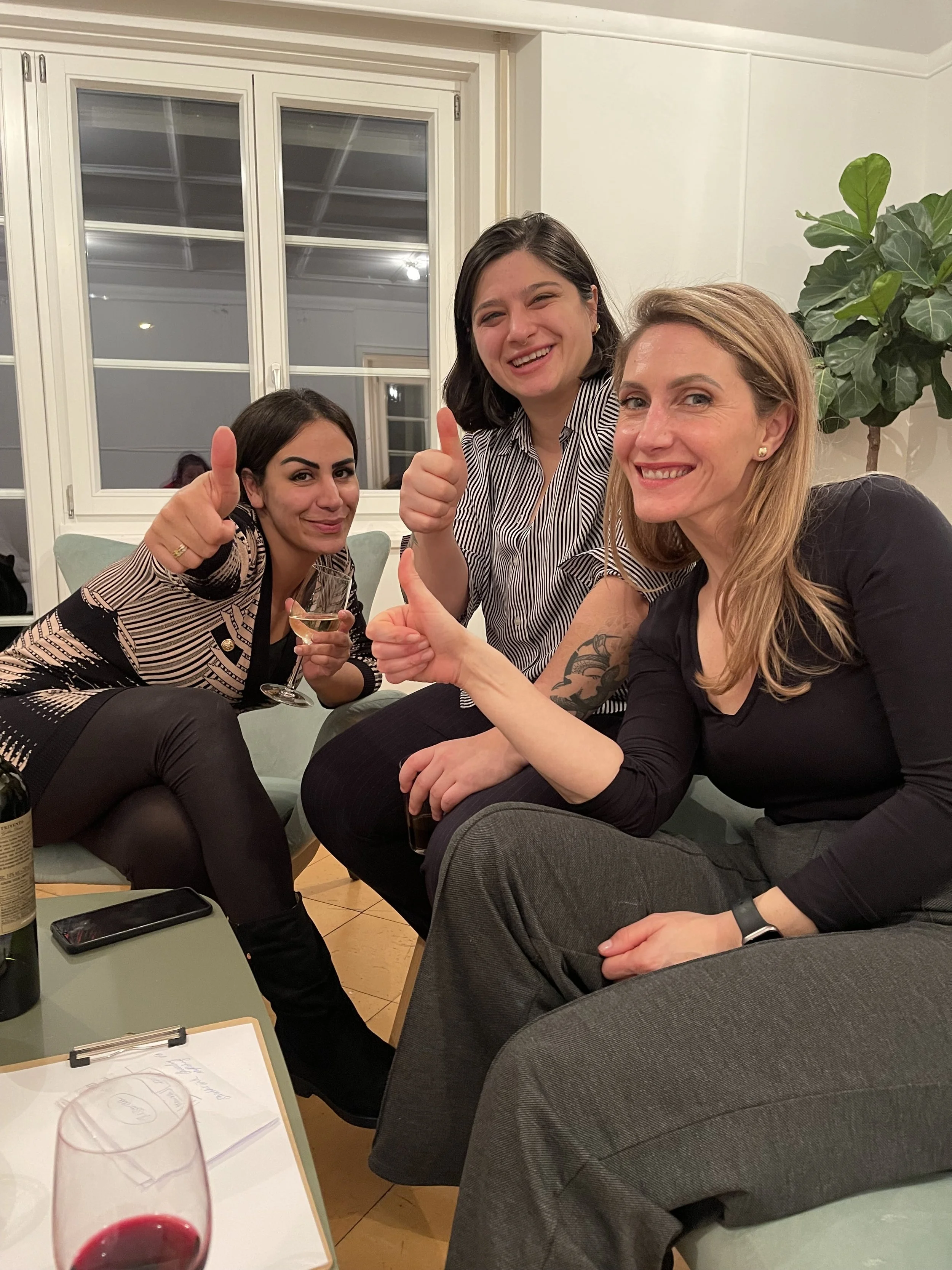 Drei Frauen sitzen auf einer Couch, lachen, geben Daumen hoch und halten Wein in einem Wohnzimmer.