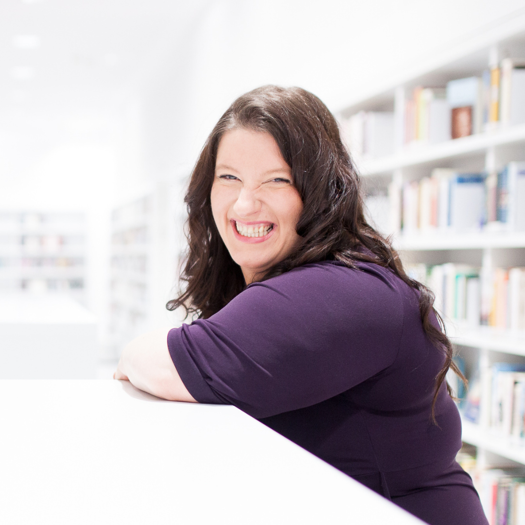 Lächelnde Frau mit langen dunklen Haaren, die auf einem weißen Tisch in einer Bibliothek sitzt.