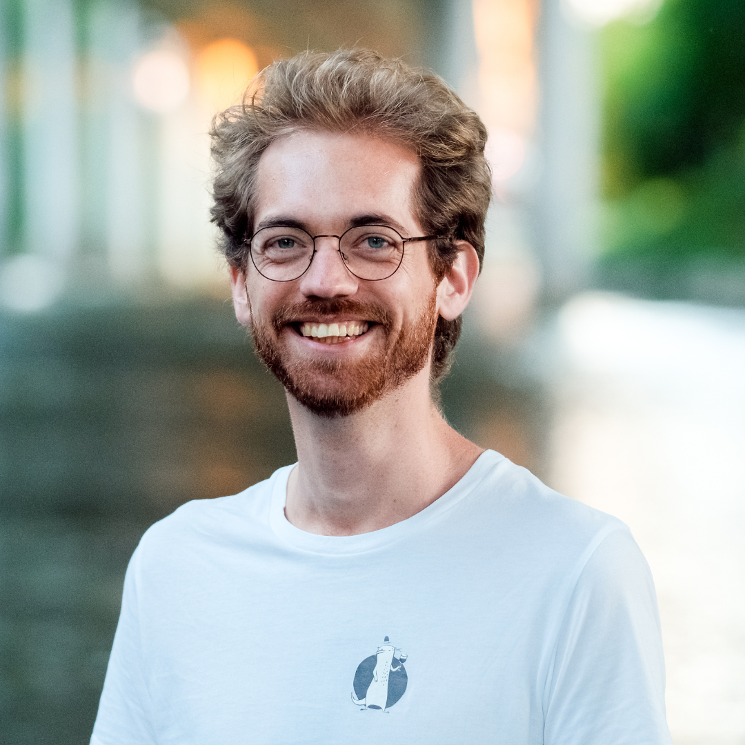 Lächelnder Mann mit Brille, hellbraunes lockiges Haar und Bart, trägt ein weißes T-Shirt, im Hintergrund unscharfe Natur mit Wasser, Bäumen und Sonnenlicht.