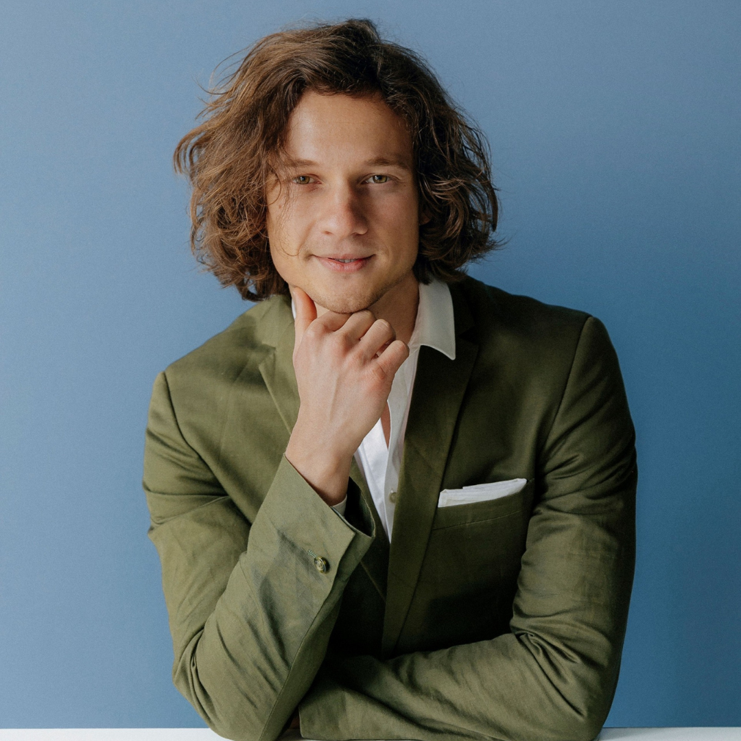 Ein junger Mann mit langen, lockigen braunen Haaren trägt einen grünen Anzug und hält die Hand an sein Kinn, vor einem blauen Hintergrund.