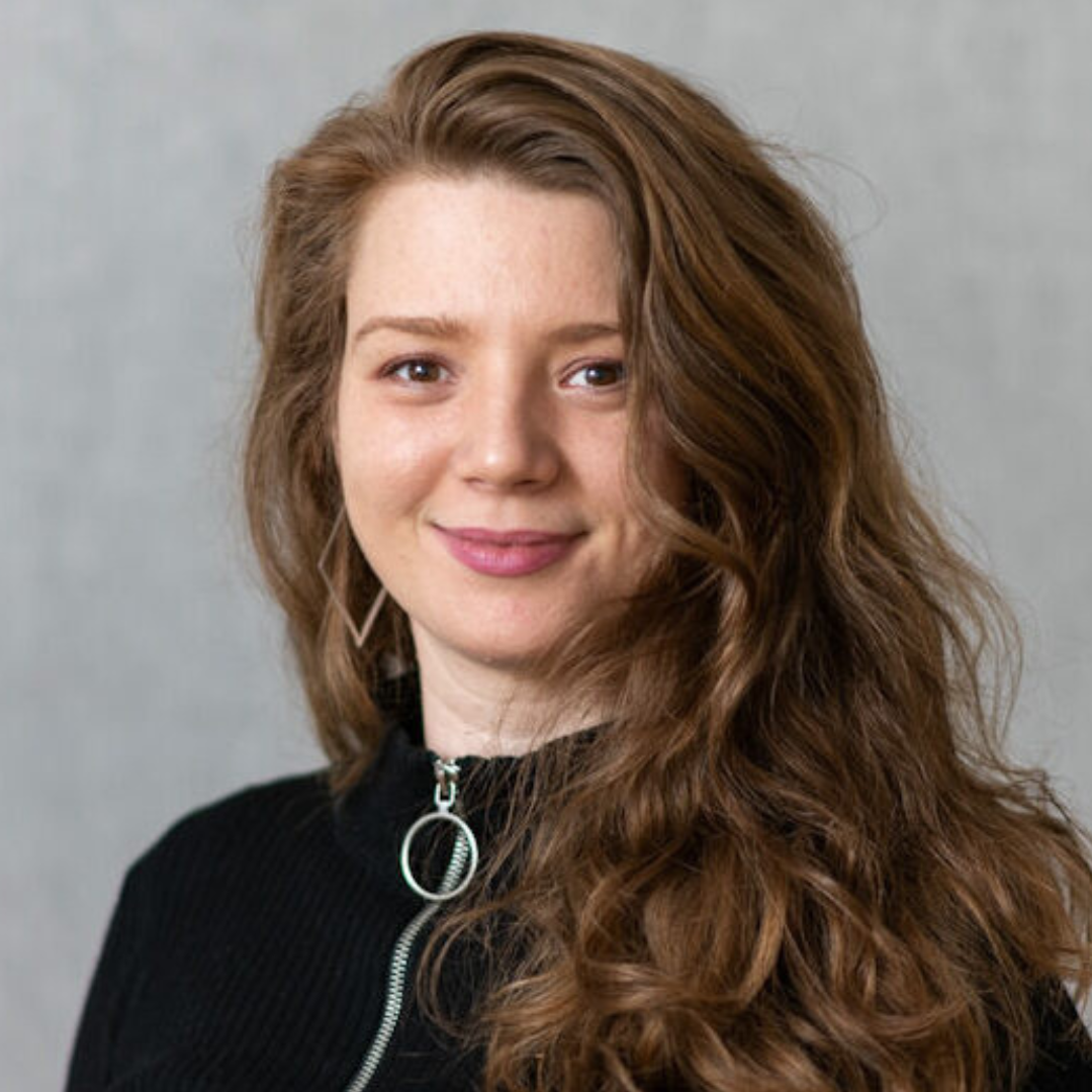 Junge Frau mit langen, lockigen braunen Haaren, trägt schwarze Jacke mit Reißverschluss, lächelt vor grauem Hintergrund.