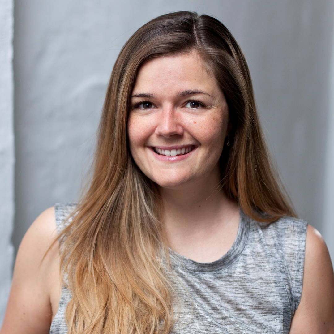 Portrait einer lachenden jungen Frau mit braunen, langem Haar, trägem grau-silbernem Oberteil, vor einem hellen Hintergrund.