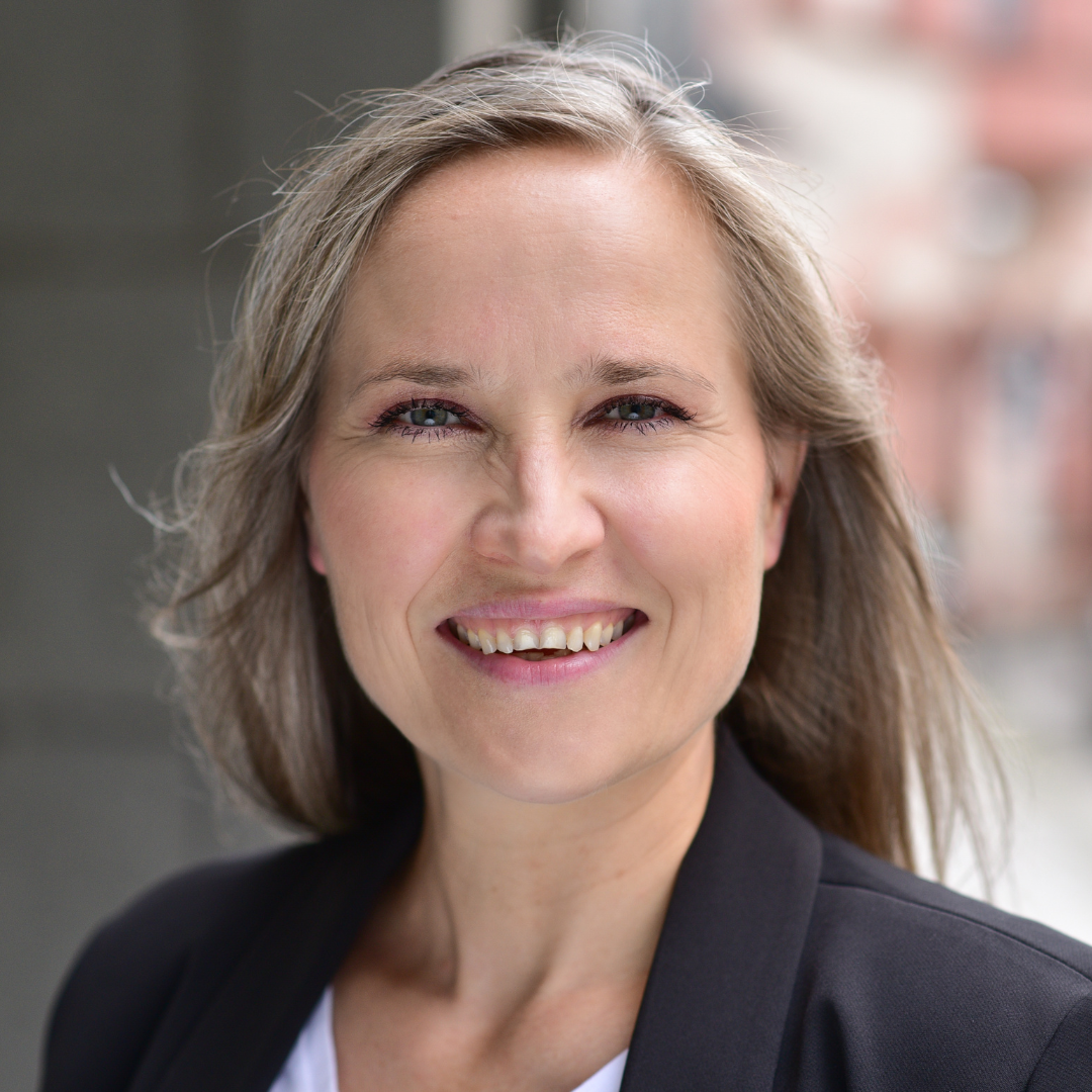 Lächelnde Frau mit blonden Haaren vor einem urbanen Hintergrund.