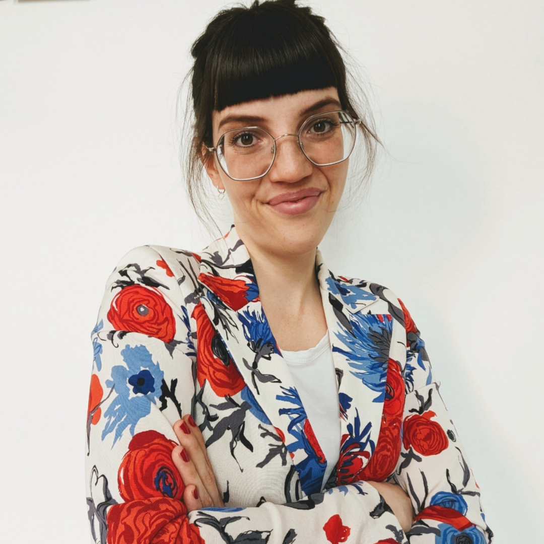 Junge Frau mit Brille, schwarzen Haaren mit Pony, trägt einen weißen Blazer mit rotem, blauem und schwarzem Blumenmuster, vor weißem Hintergrund.