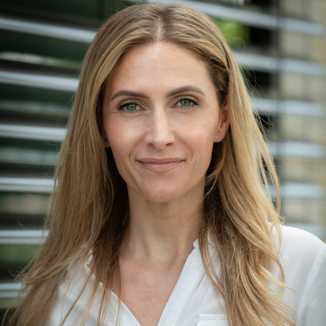 Nahaufnahme einer lachenden Frau mit langem, blondem Haar, tragend weißes Hemd, vor einem modernen Gebäude mit horizontalen Fensterläden.