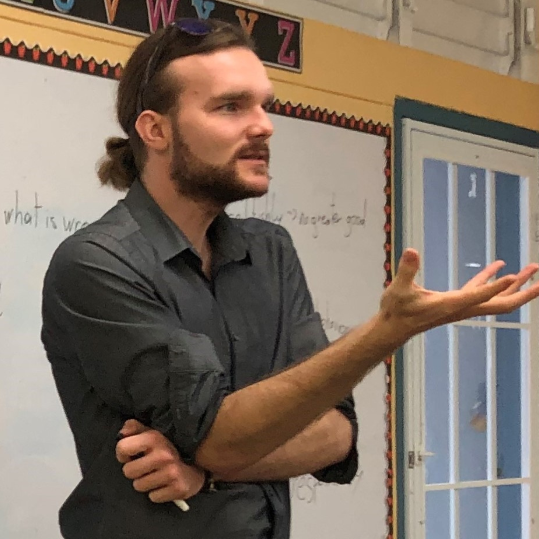 Ein Mann mit braunem Haar, Bart und Sonnenbrille auf dem Kopf steht in einem Klassenzimmer und erklärt etwas. Im Hintergrund sind Buchstaben an der Wand und eine Tafel.