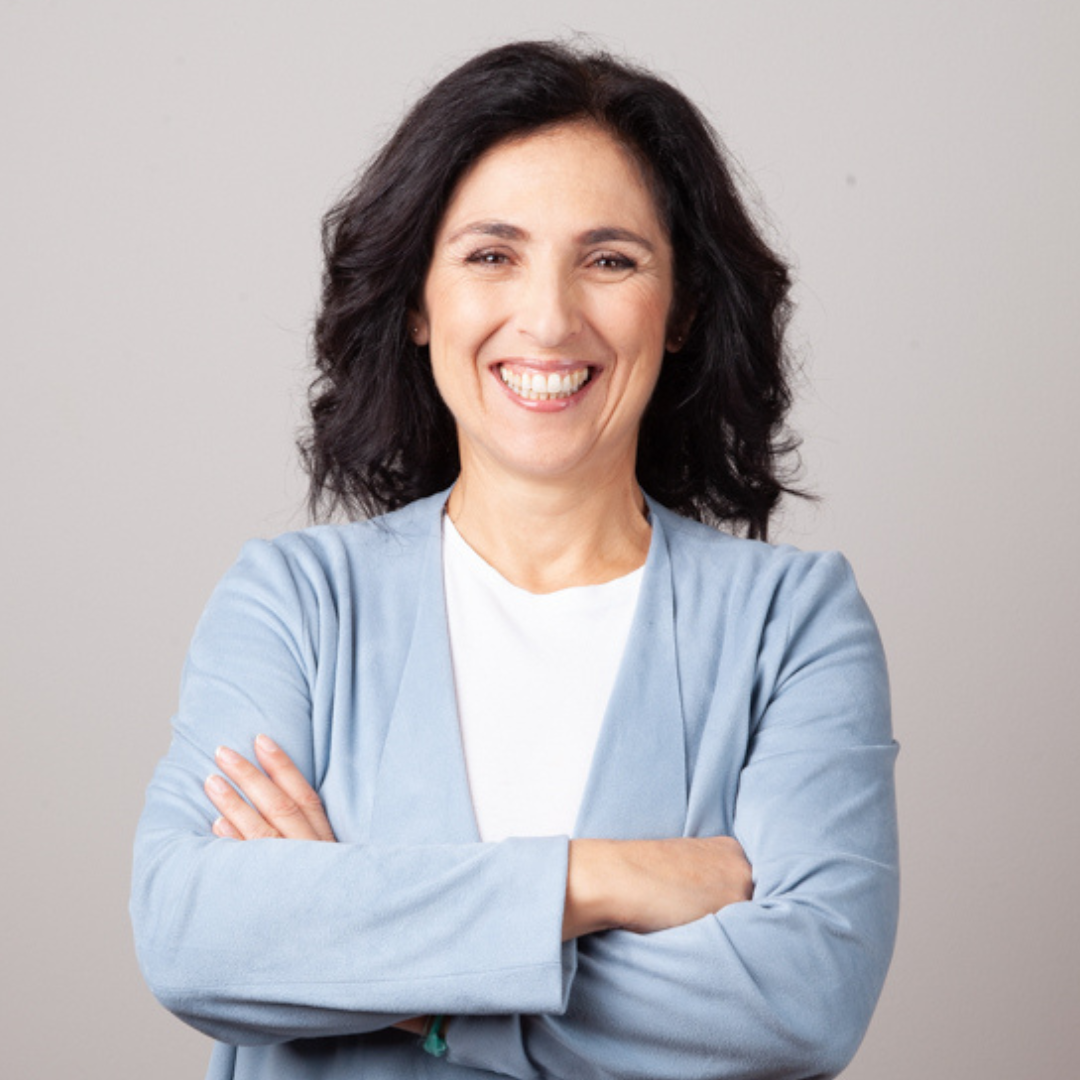 Eine lachende Frau mit schwarzen, lockigen Haaren trägt eine blaue Jacke über einem weißen Shirt, steht vor einem grauen Hintergrund, Arme verschränkt.