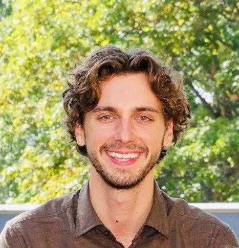 Ein junger Mann mit braunen, gewellten Haaren und einem Bart, der vor einer grünen, blättrigen Baumkulisse lächelnd in die Kamera schaut.