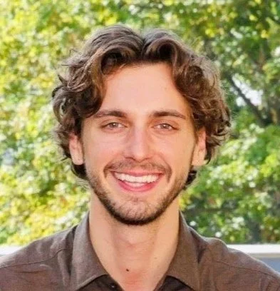 Ein junger Mann mit braunen, lockigen Haaren, Bart und braunen Augen lächelnd vor einem grünen Baum im Freien.