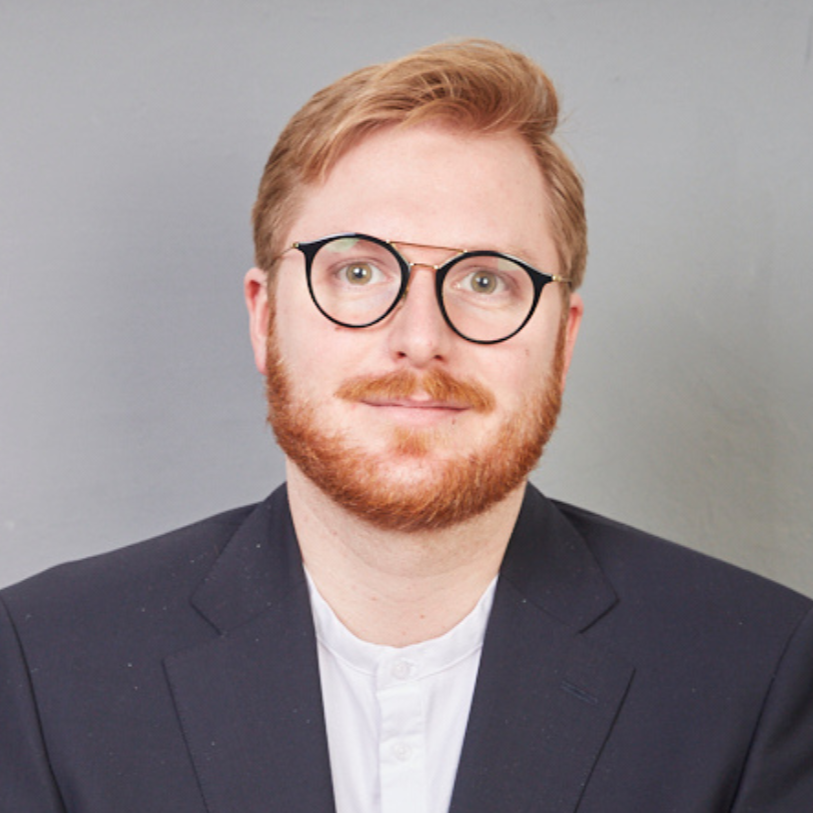 Porträt eines Mannes mit rotem Haar, Brille und Bart, trägt einen dunklen Anzug und ein weißes Hemd, vor grauem Hintergrund.