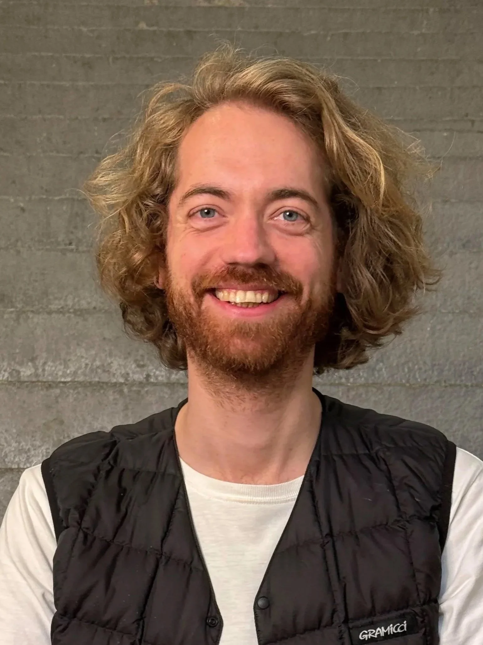 Ein Lächelnder Mann mit lockigem, braunem Haar und einem Bart, trägt ein weißes T-Shirt und eine schwarze Weste, vor einem grauen Betonhintergrund.