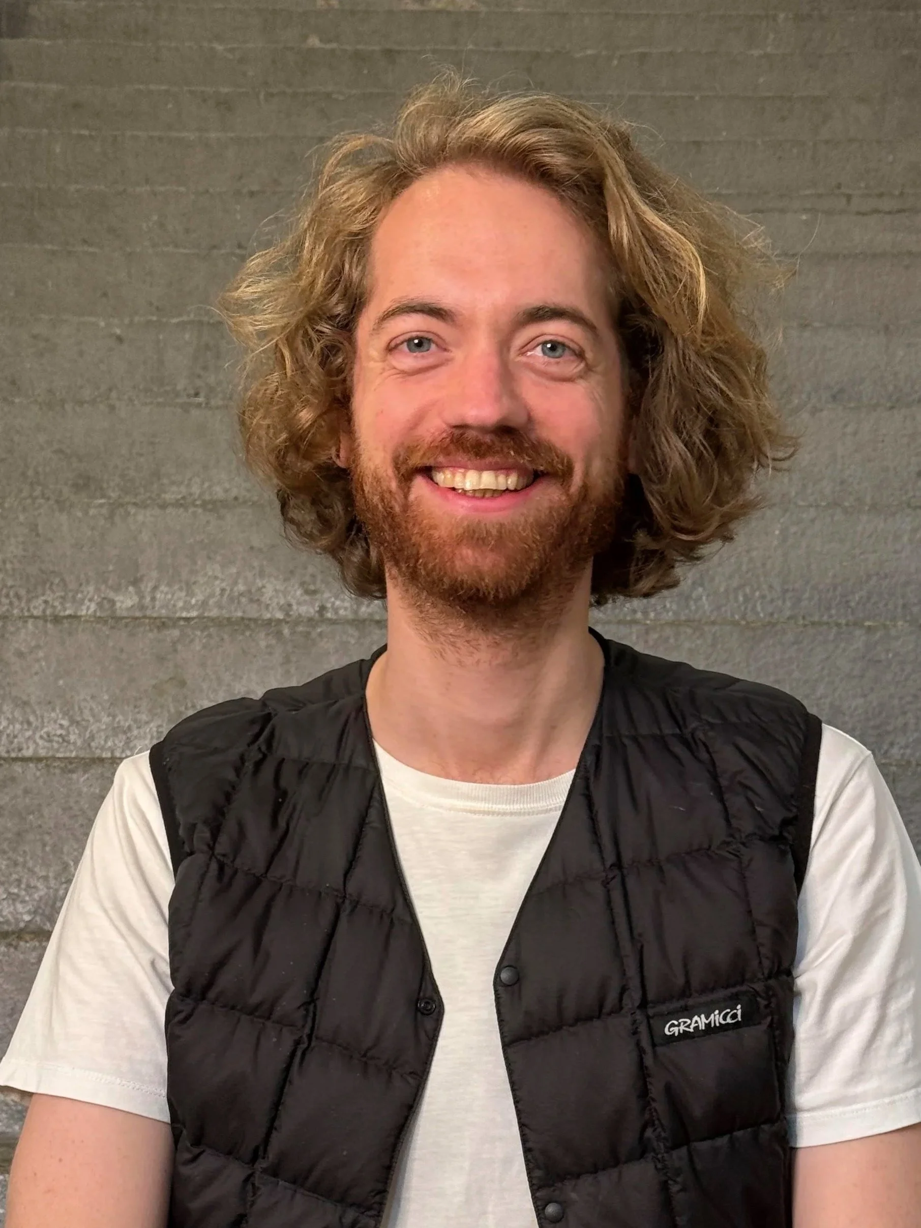 Ein Mann mit lockigem, braunem Haar und Bart, trägt ein weißes T-Shirt und eine schwarze, wattierte Weste, steht vor einer grauen Steinumwall.