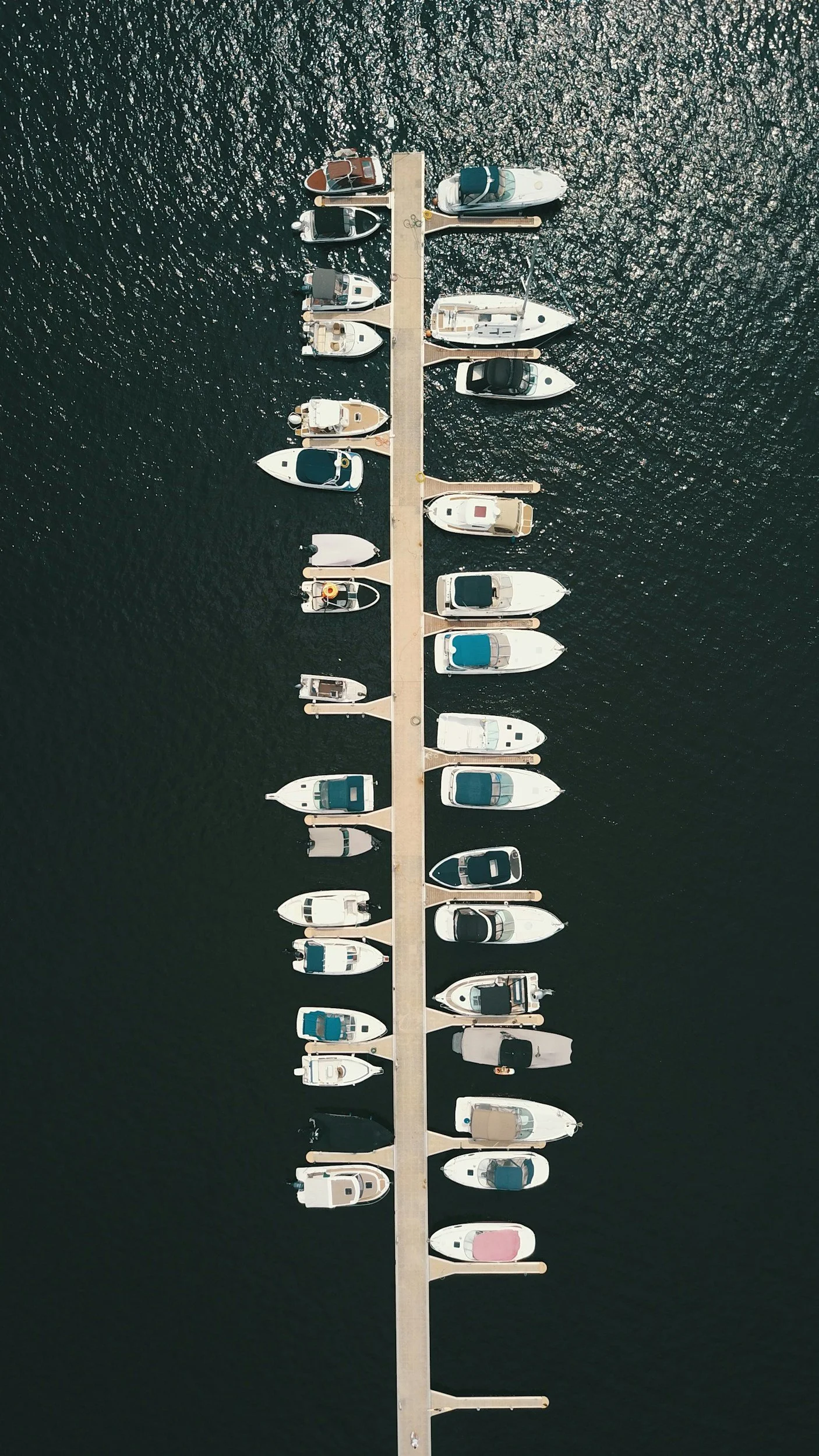 An aerial view of a marina with multiple boats docked along a long pier extending into dark water.