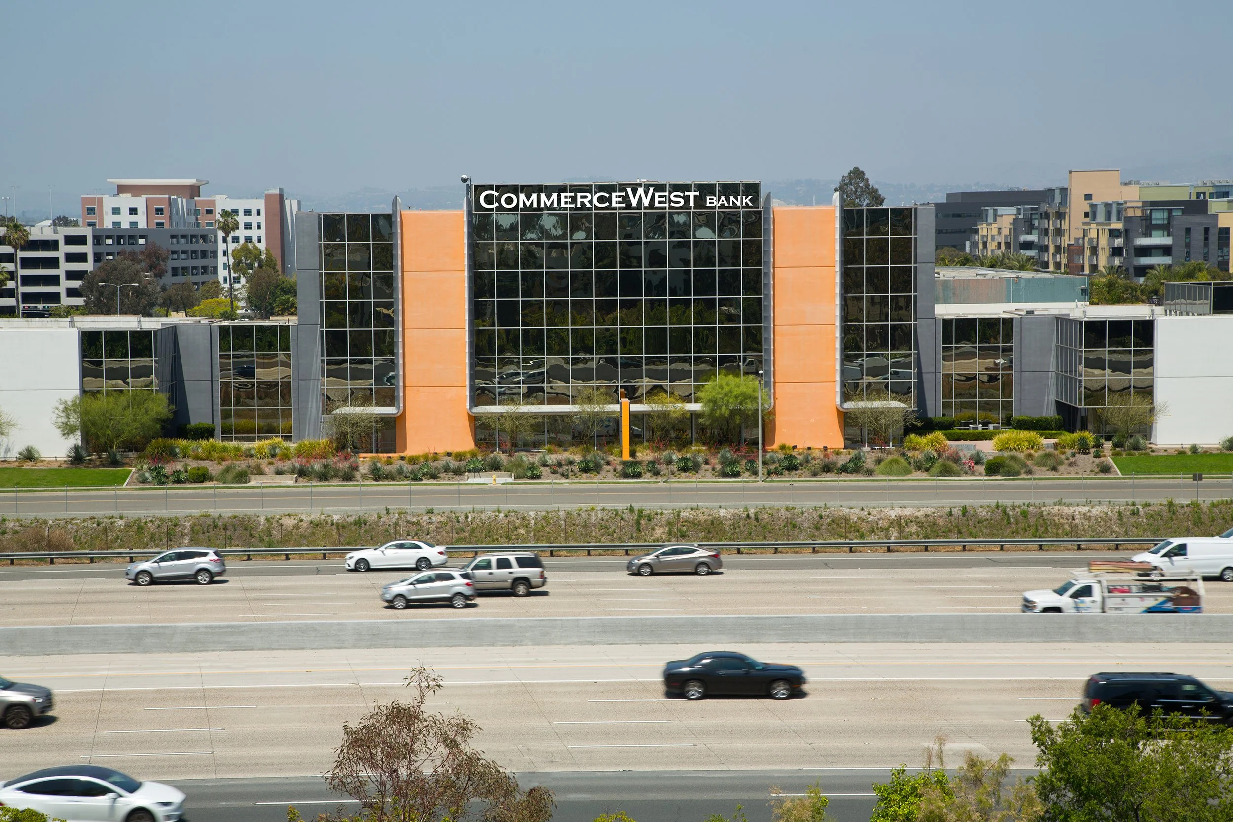 A modern office building labeled 'CommerceWest Bank' with glass windows and landscaped greenery in front, visible from a highway with passing cars.